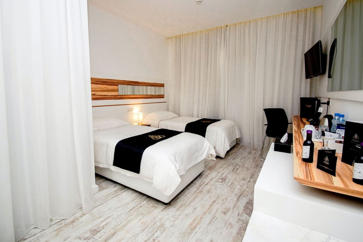 Une chambre d’hôtel avec deux lits, un bureau équipé d’un ordinateur et de bouteilles de vin, et un téléviseur fixé au mur.