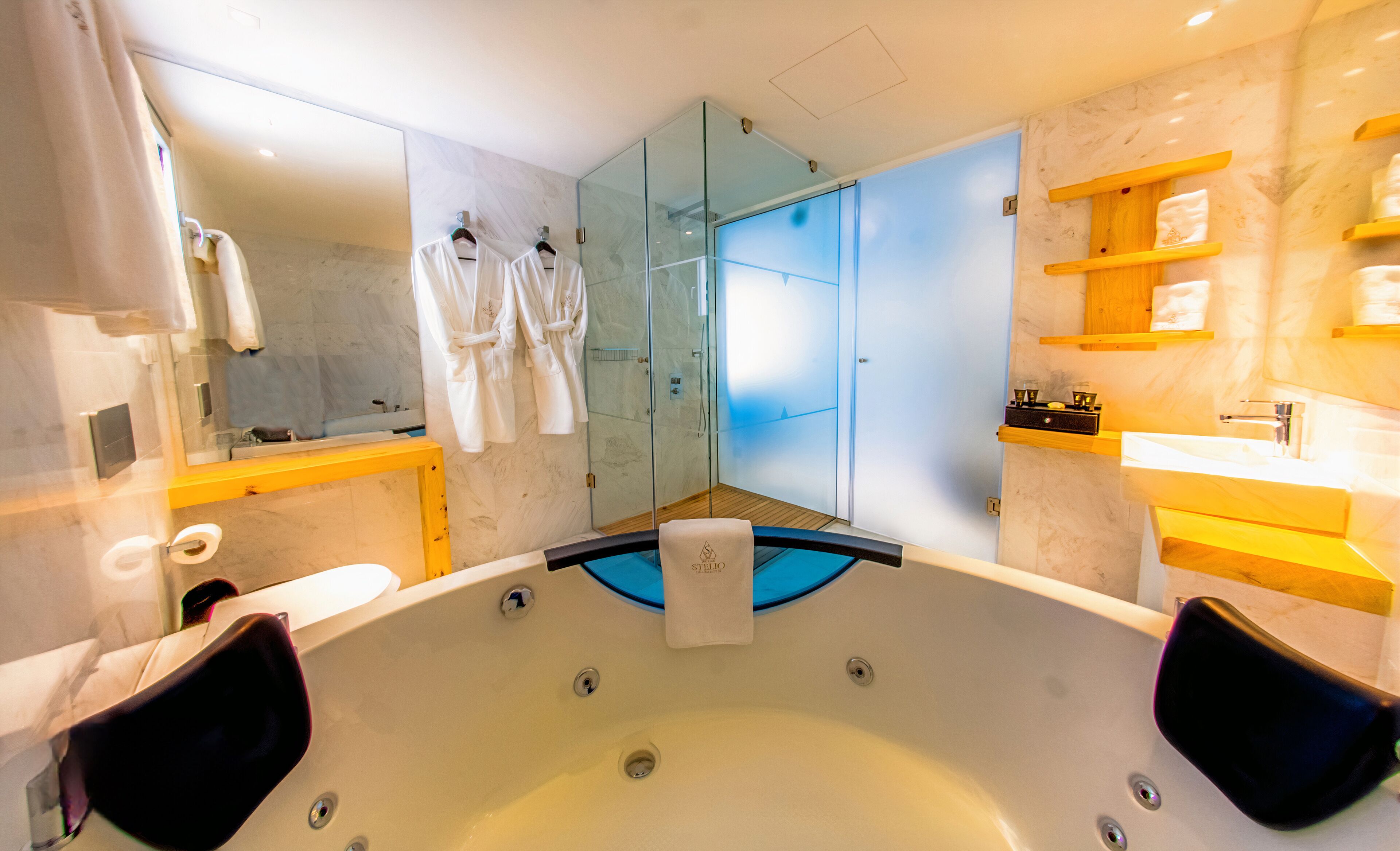 Un baño moderno con ducha de vidrio, una bañera grande con dos sillas y un tocador de madera con estantes.
