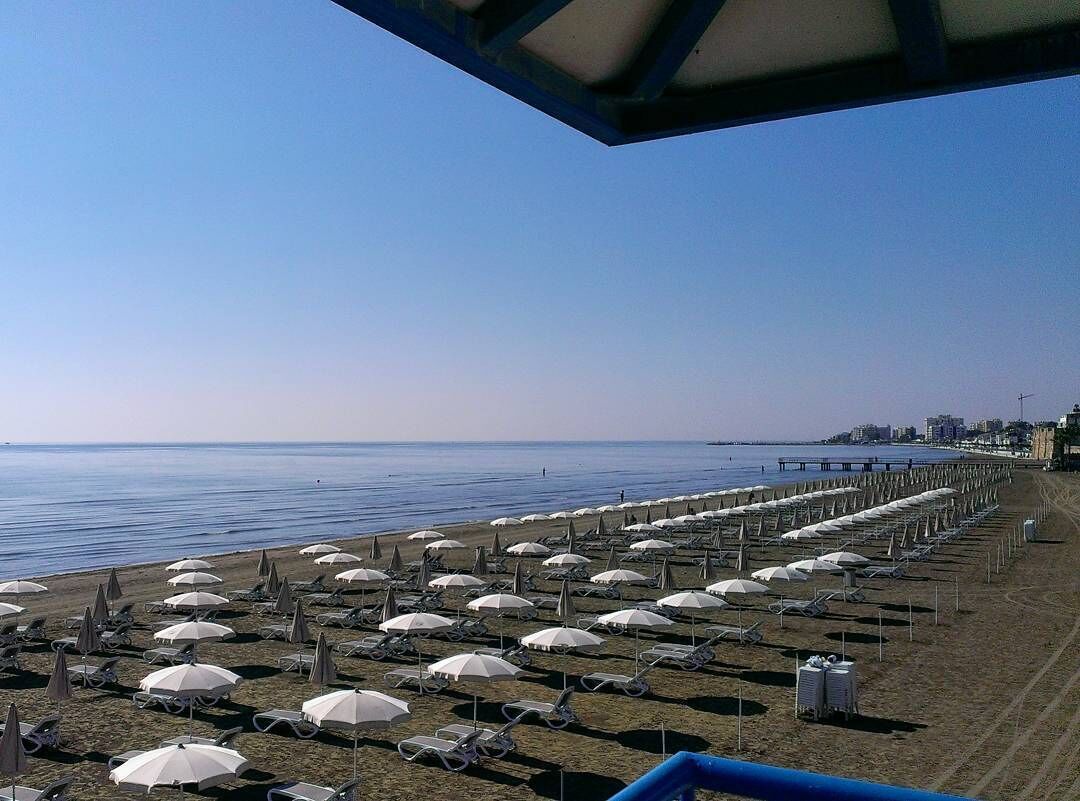 Una playa con hileras de sombrillas y sillas de descanso, un muelle al fondo y un cielo despejado.
