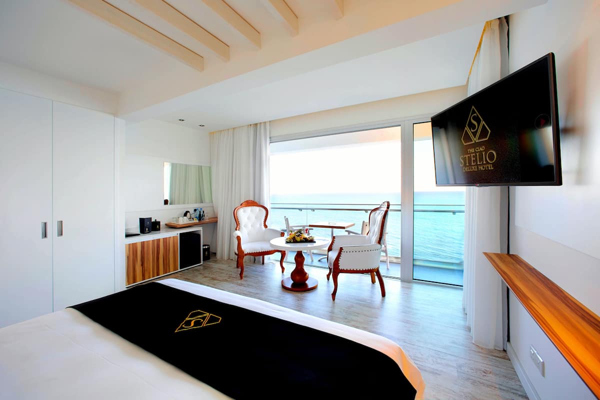 Une chambre d’hôtel moderne équipée d’un lit, d’une télévision, d’un balcon avec vue sur la mer et d’un coin repas avec des chaises et une table.