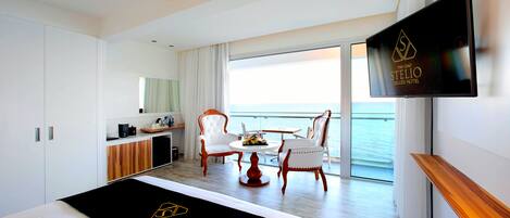 A modern hotel room with a bed, a TV, a balcony with a view of the sea, and a dining area with chairs and a table.