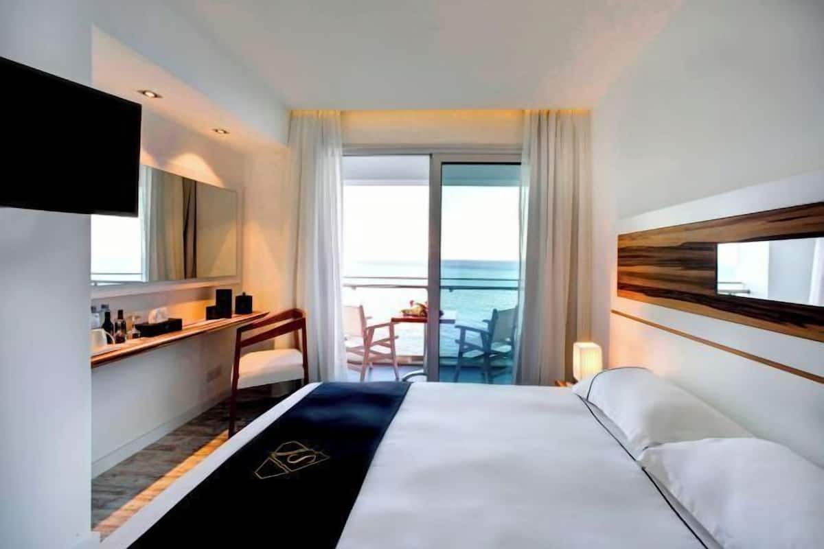 Une chambre d’hôtel comprenant un lit, un bureau, une chaise, une télévision, un balcon avec vue sur la mer et une porte coulissante.
