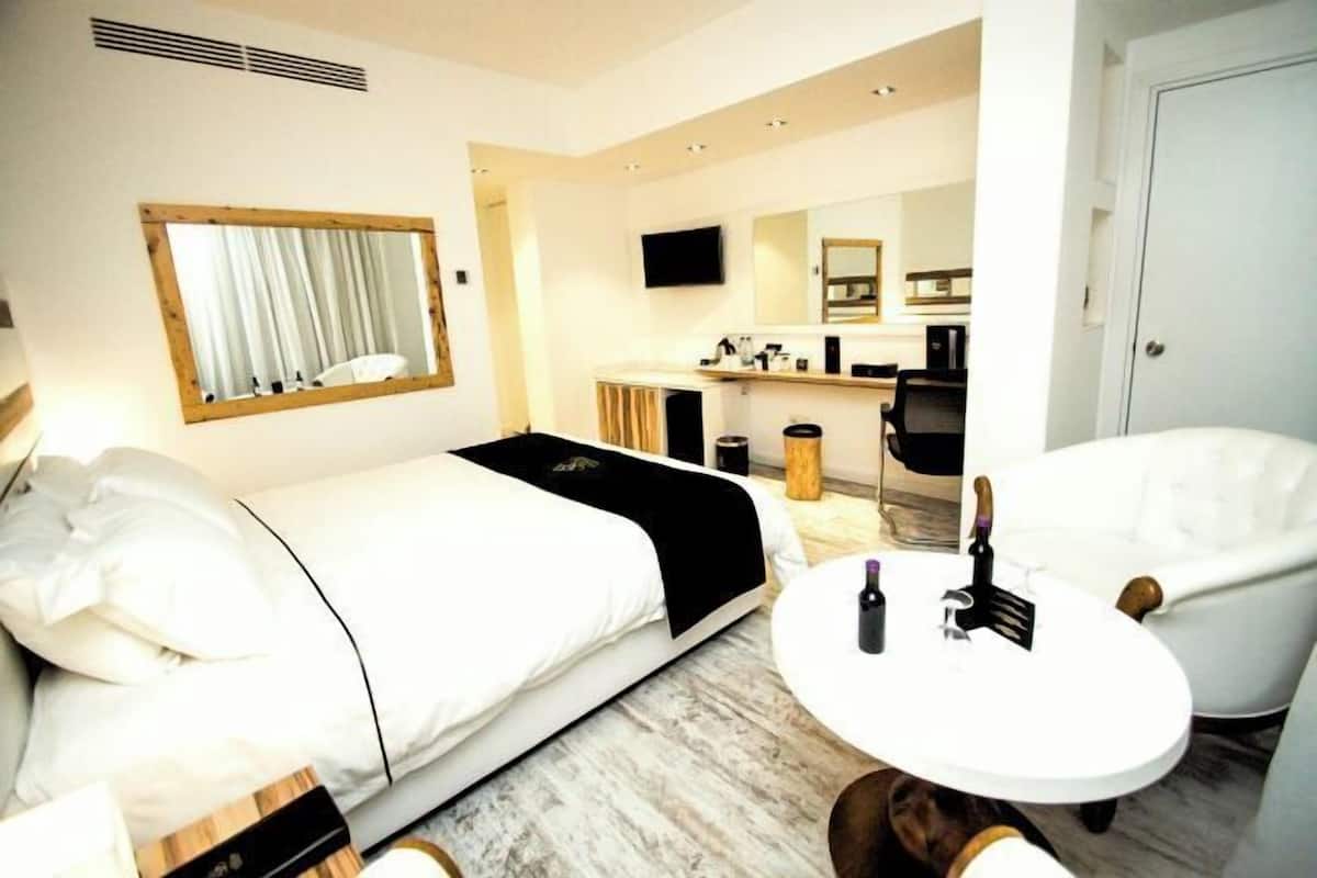 Une chambre d’hôtel avec un lit, une table ronde sur laquelle se trouvent une bouteille et une fourchette, un fauteuil blanc, un miroir et, au fond, une petite cuisine.