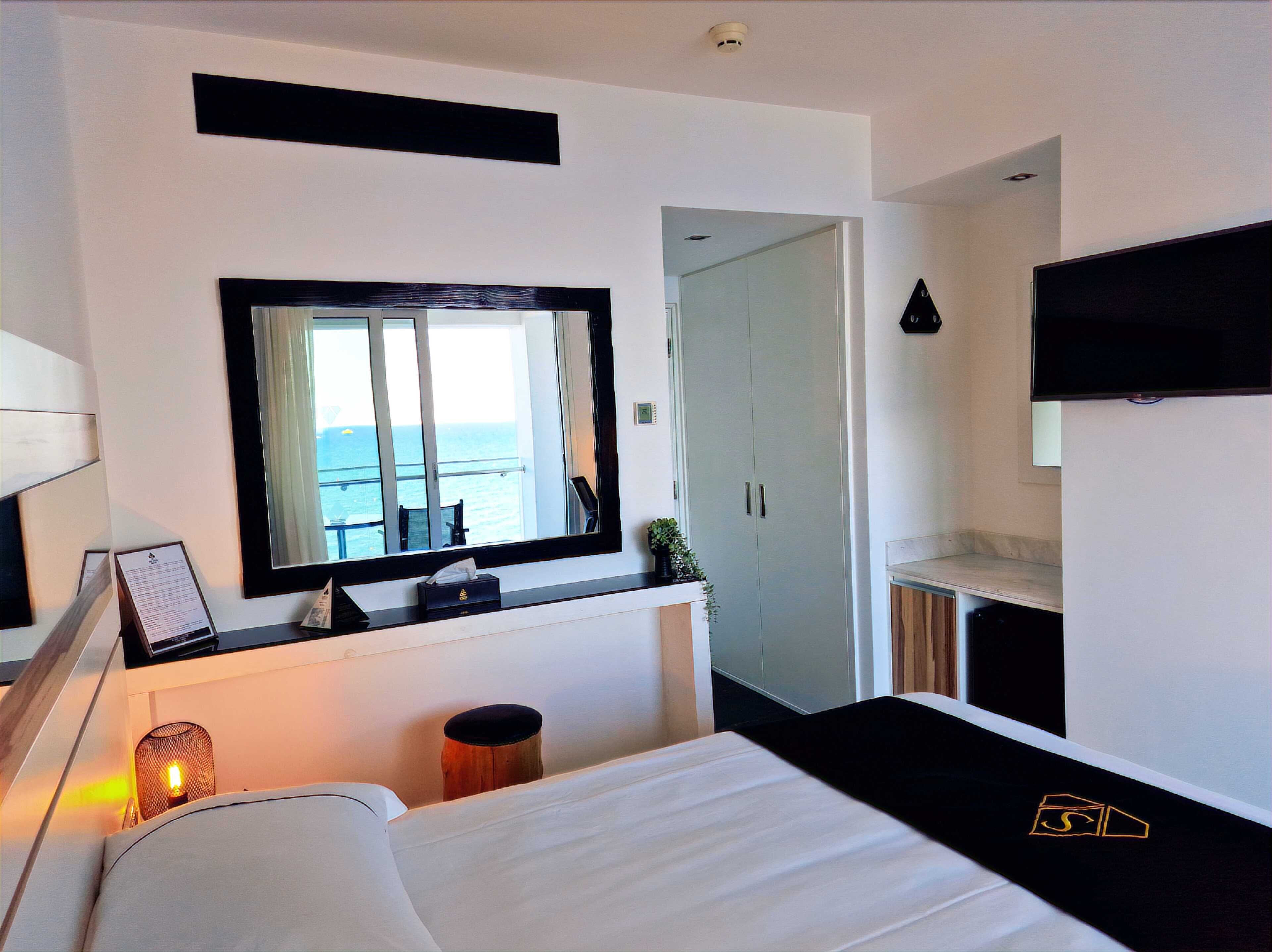 A hotel room with a bed, a large mirror, a television, a wardrobe, and a view of the sea.