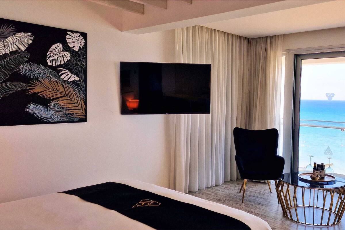 Une chambre d’hôtel avec un lit, une télévision, deux fauteuils, une petite table avec des boissons et un balcon donnant sur la mer.
