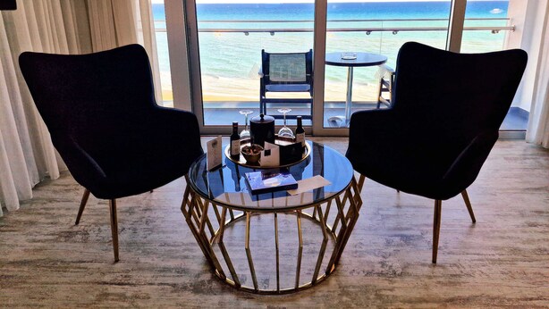 Une pièce avec deux chaises noires, disposées face à une table en verre sur laquelle se trouve un plateau d’objets. De grandes fenêtres offrent une vue sur une plage et l’océan.