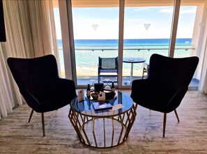 Une pièce avec deux chaises noires, disposées face à une table en verre sur laquelle se trouve un plateau d’objets. De grandes fenêtres offrent une vue sur une plage et l’océan.