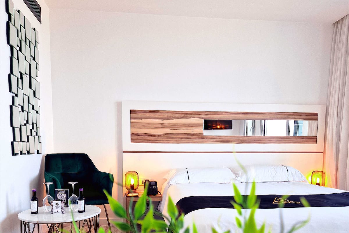 Une chambre d’hôtel avec un lit, des lampes de chevet, une petite table avec des bouteilles de vin, un fauteuil vert et une œuvre d’art accrochée au mur.