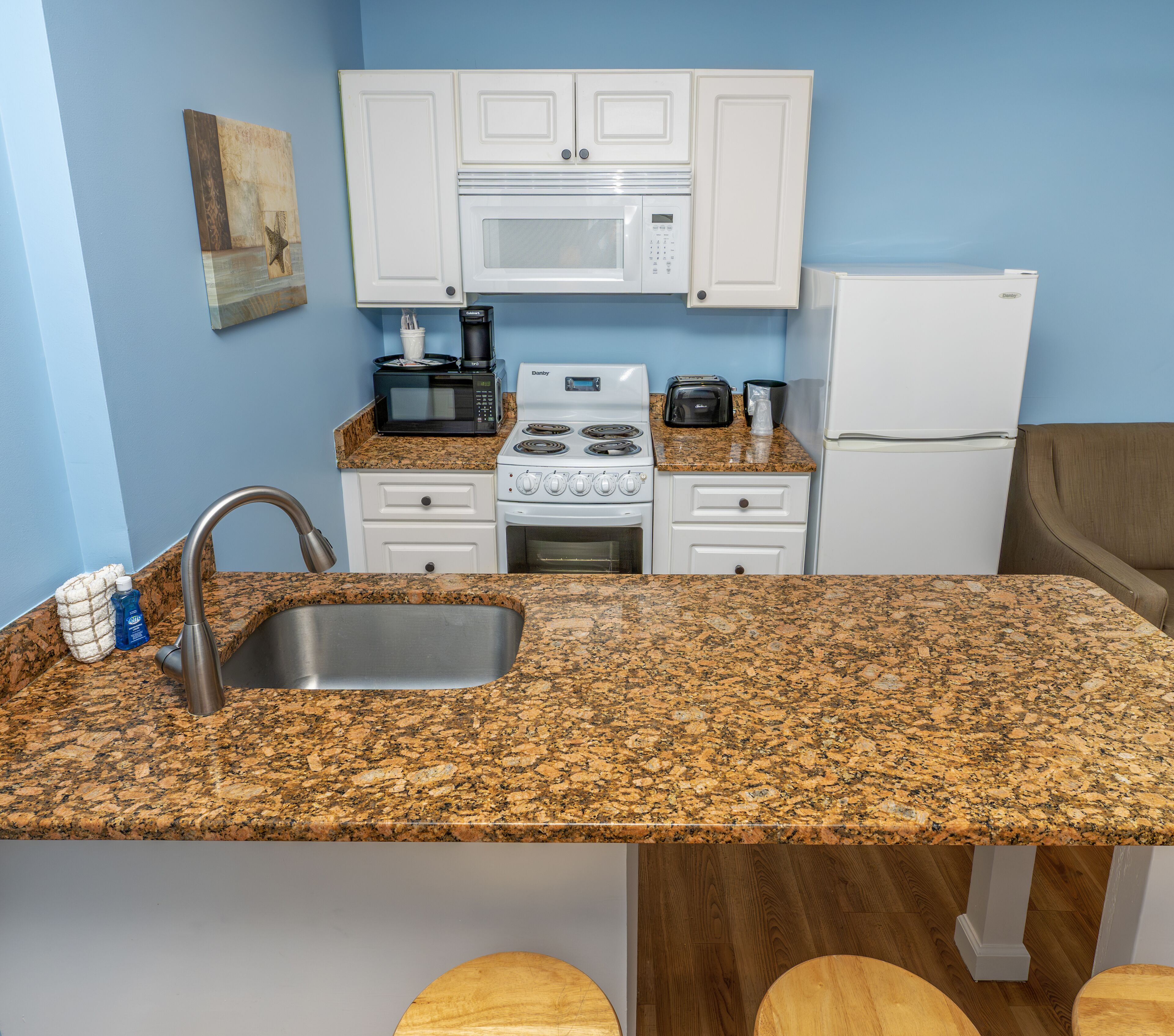 A compact kitchen with white cabinets, a granite countertop, a sink, a stove, a microwave, a toaster, and a refrigerator.