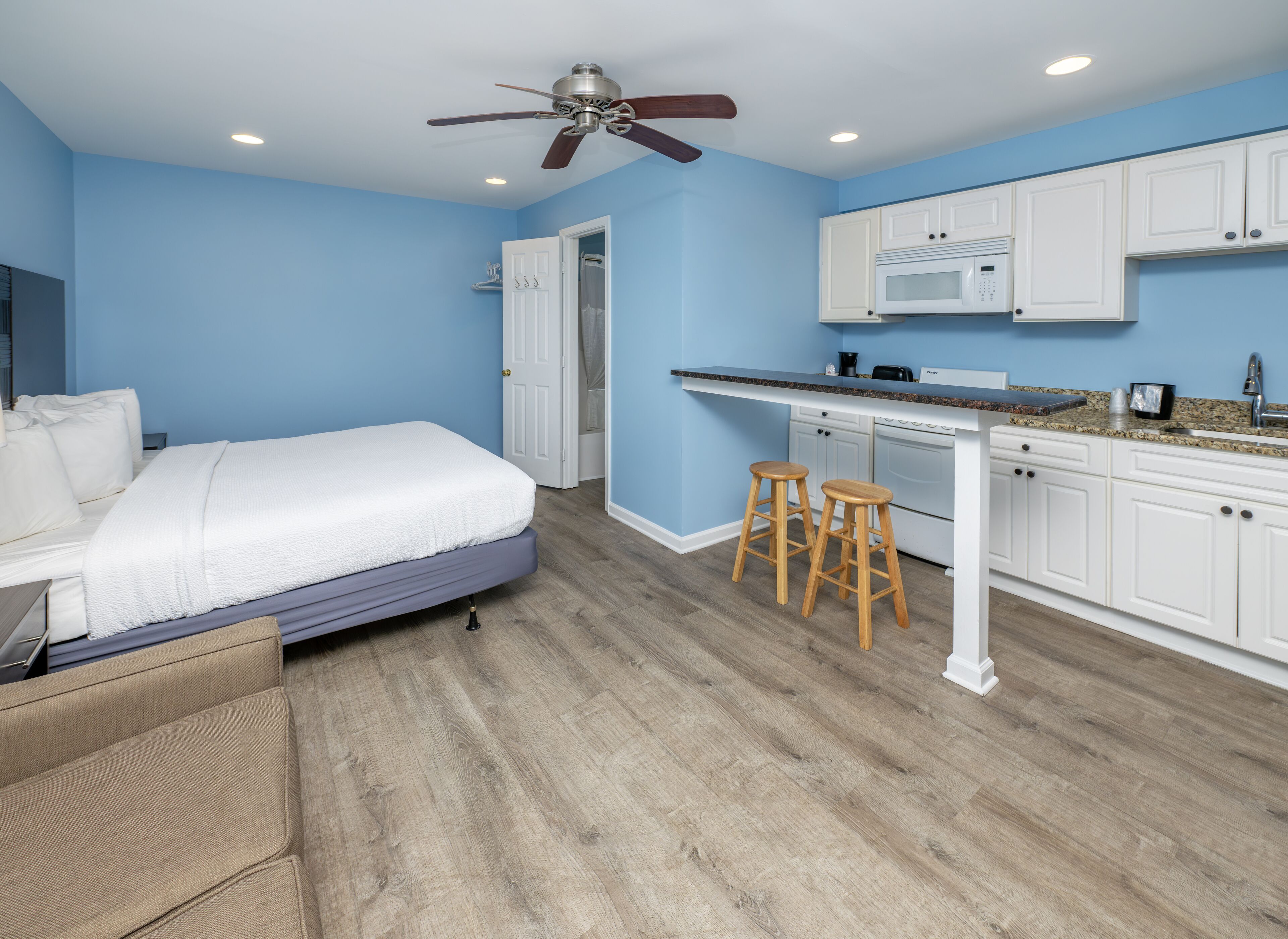 A bedroom with a bed, a ceiling fan, a kitchenette with a microwave, and a door leading to another room.