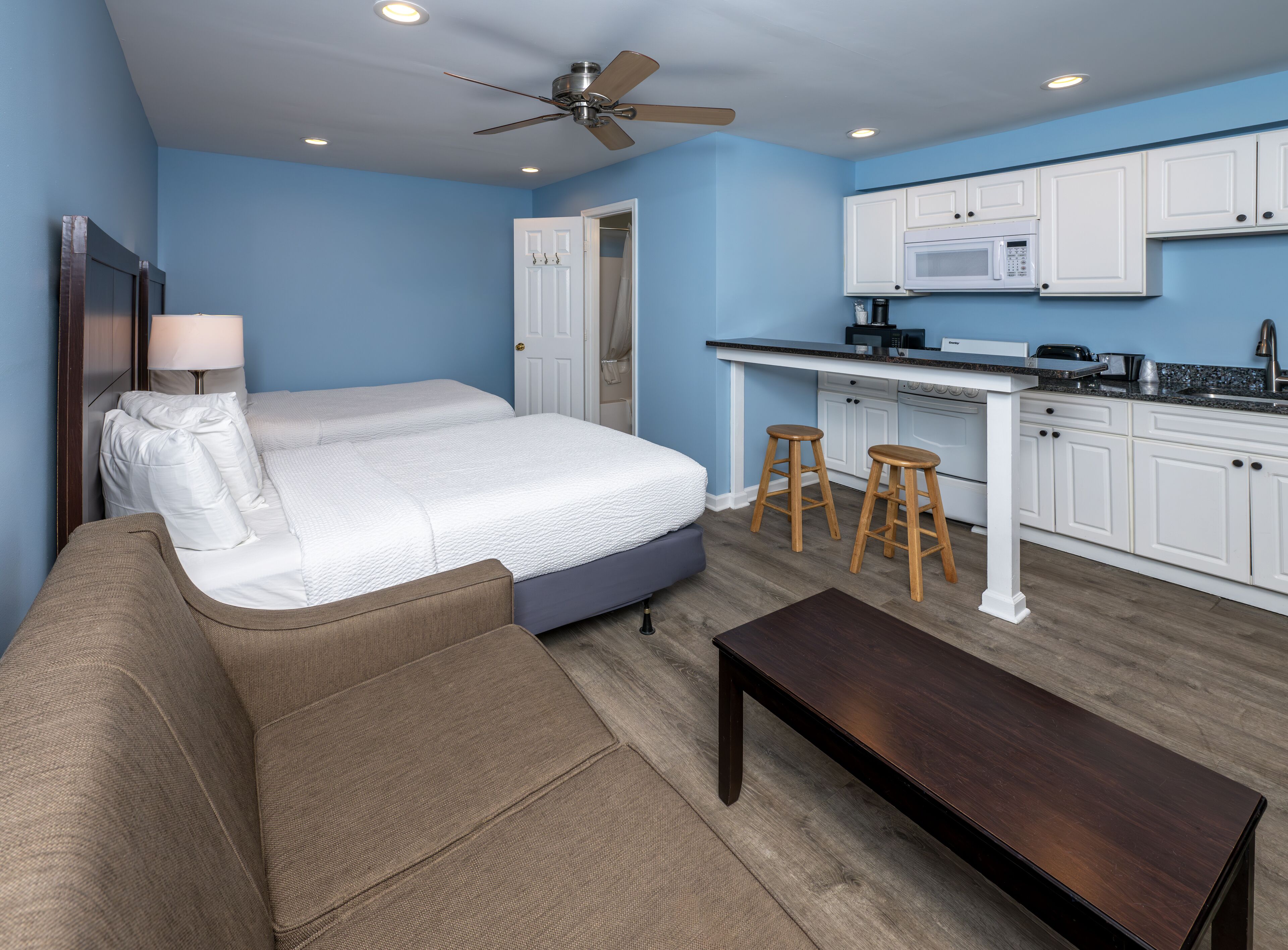 A compact room with a bed, a sofa, a small table, a kitchen area with a microwave, and a ceiling fan.