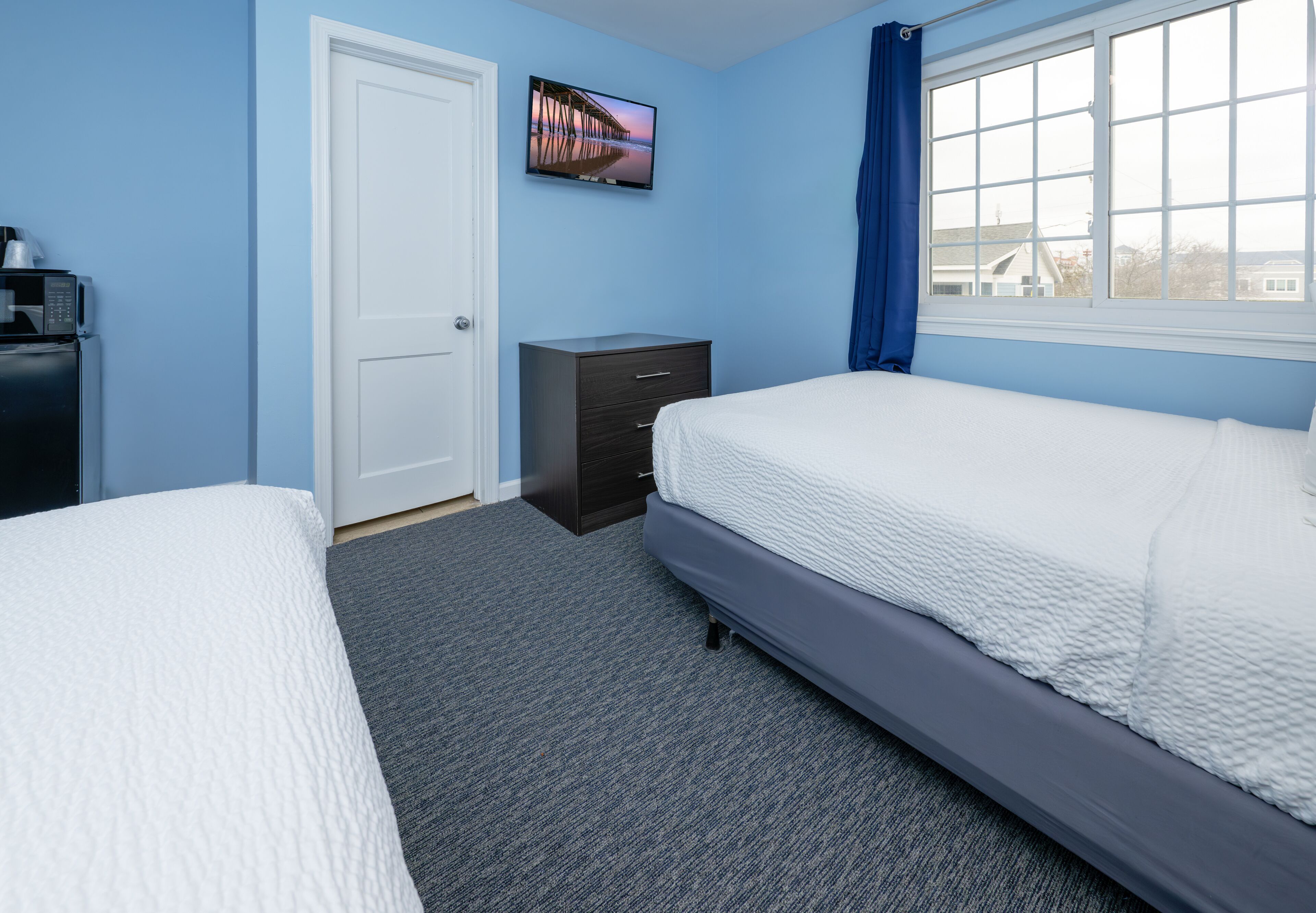 A bedroom with two beds, a TV on the wall, a dresser, and a window with curtains.