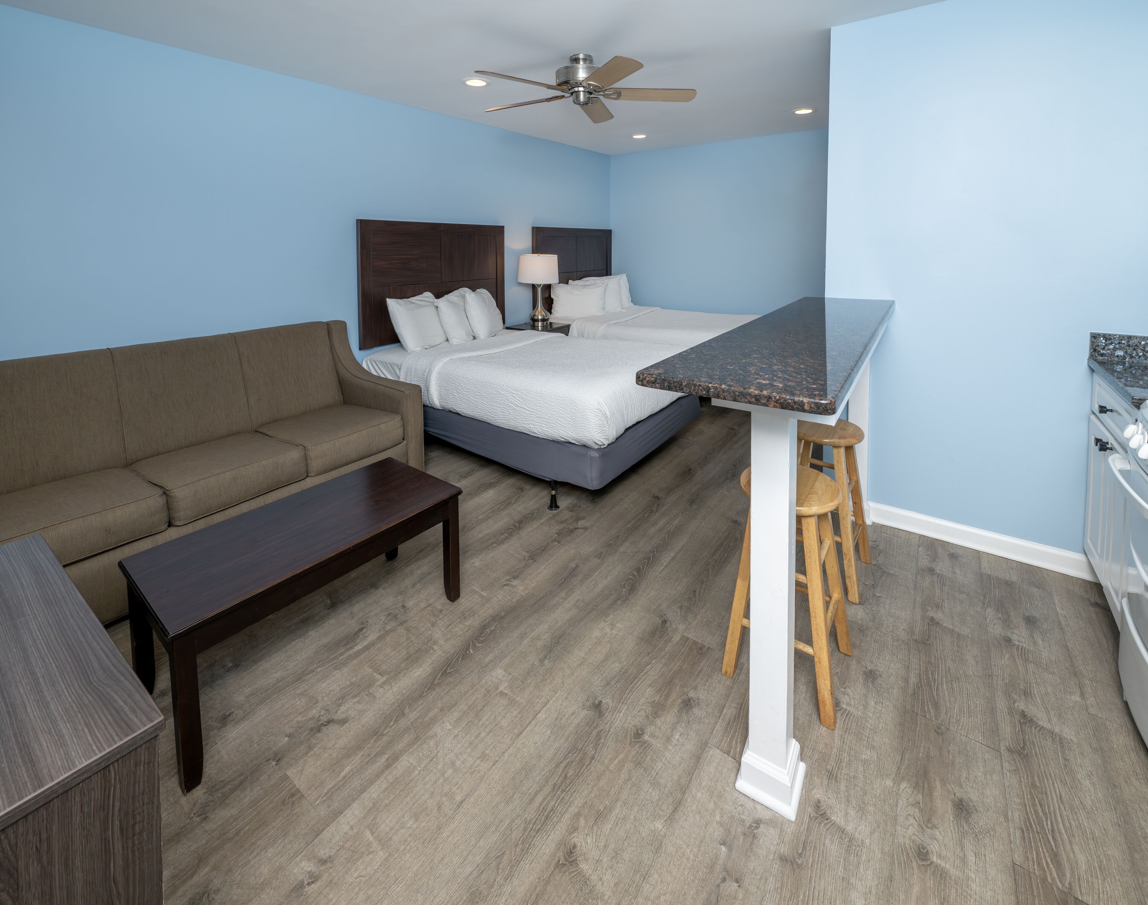 A compact hotel room with a bed, a sofa, a small table, a bar area with stools, and a ceiling fan.