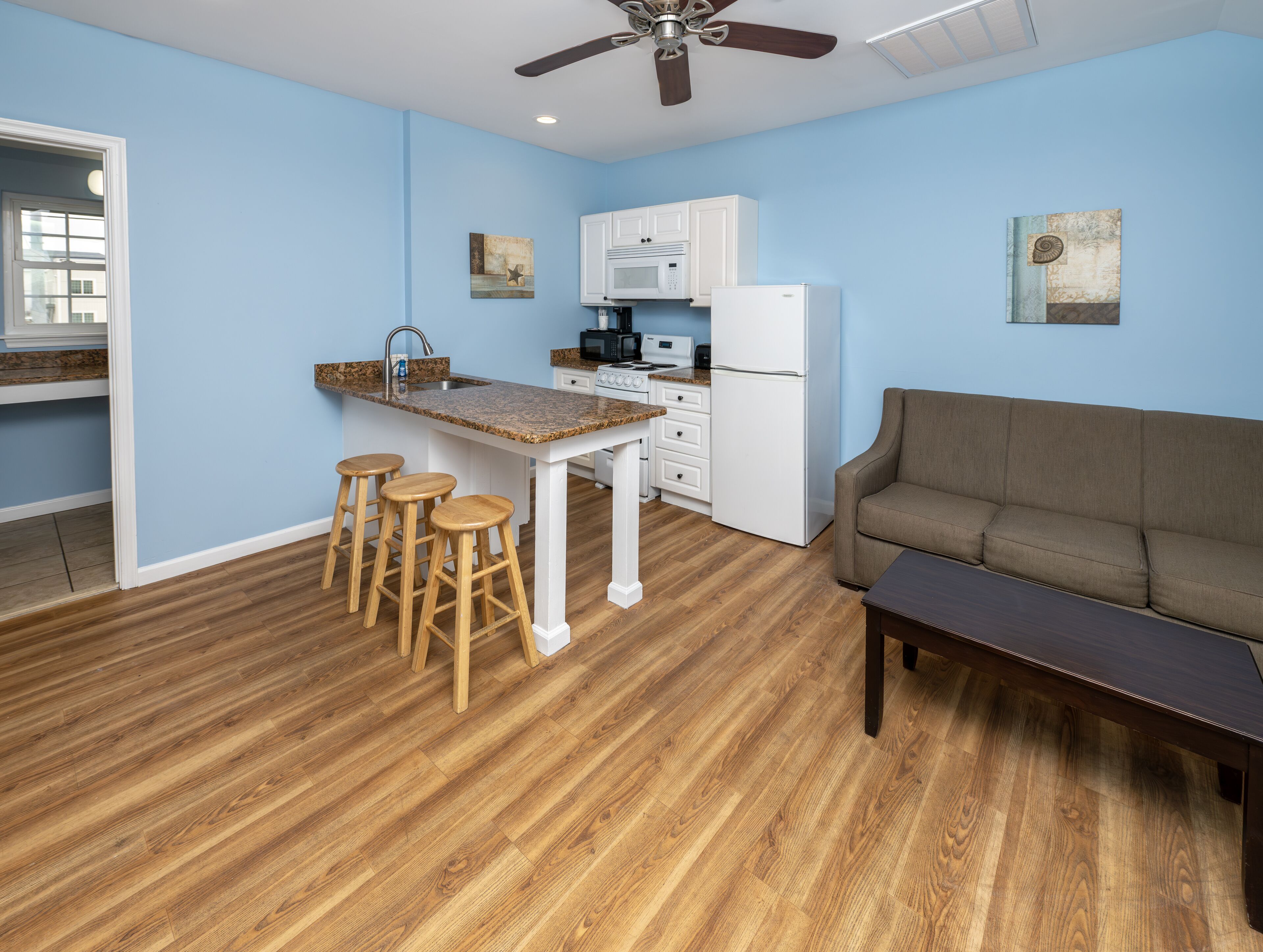 A modern kitchen with a central island, wooden stools, a white refrigerator, and a ceiling fan.