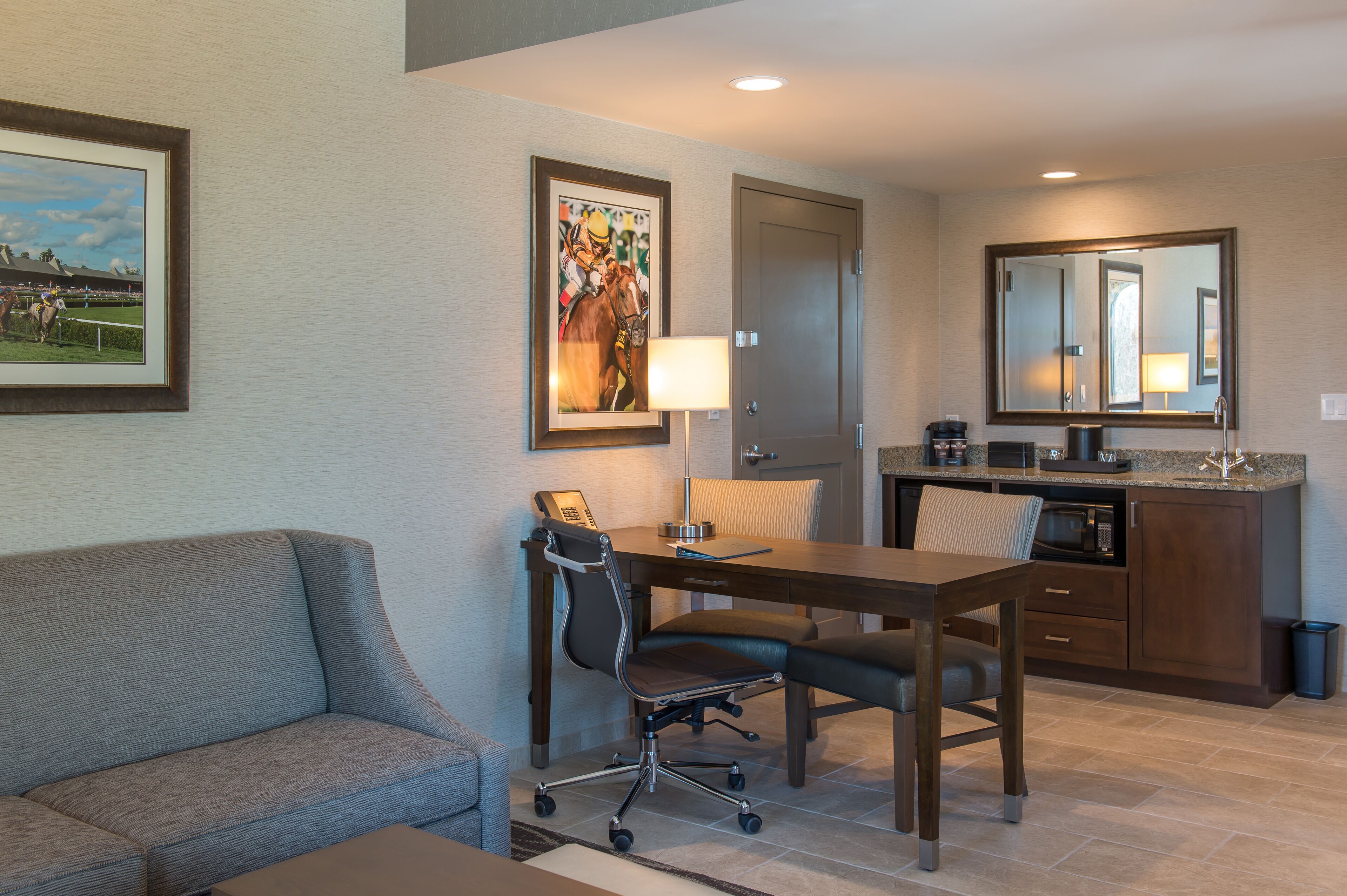 A hotel room with a sofa, a desk with a chair, a kitchen area, and a door.