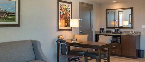 A hotel room with a sofa, a desk with a chair, a kitchen area, and a door.