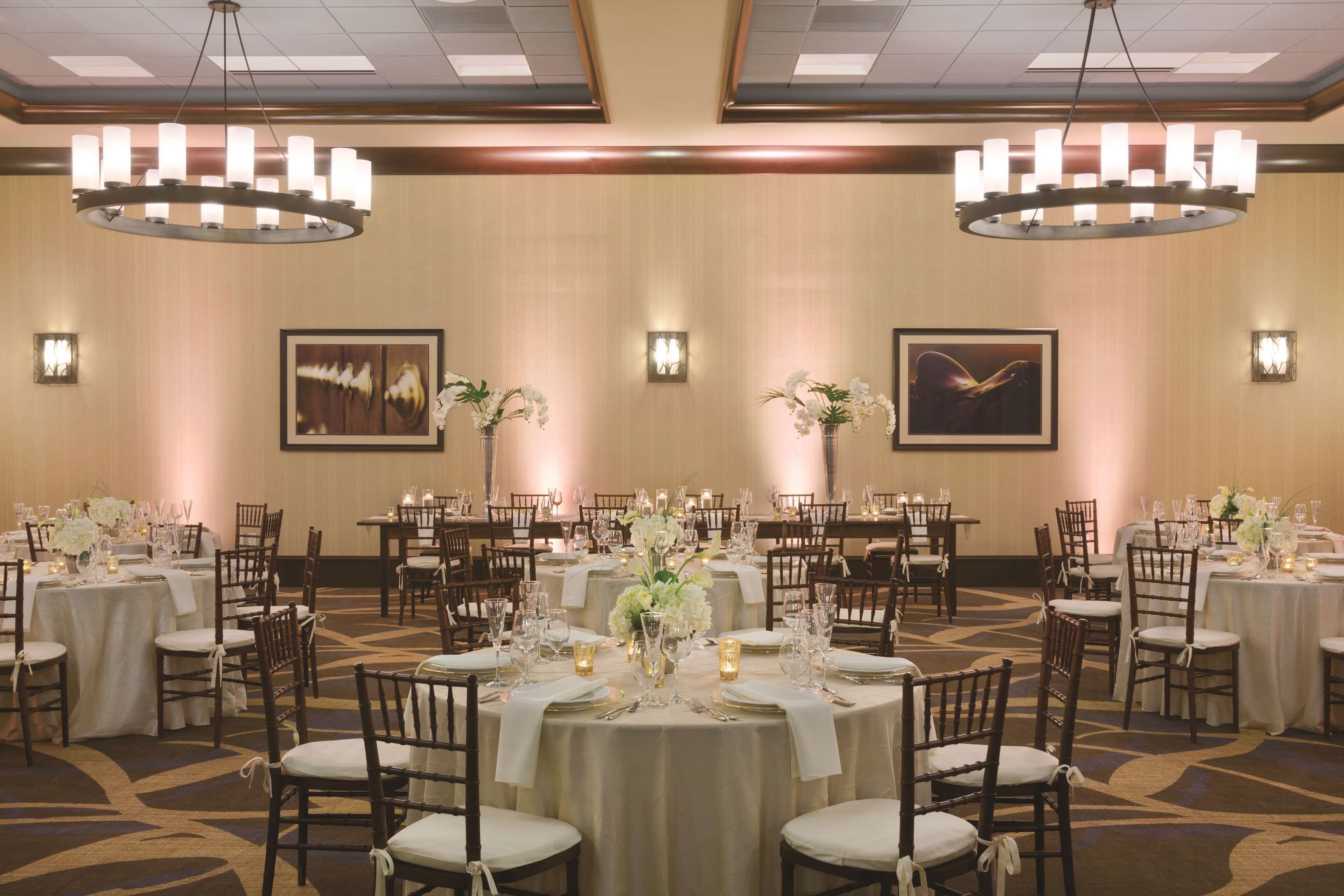 A banquet hall with round tables set for events, featuring chandeliers, floral centerpieces, and framed artwork on the walls.