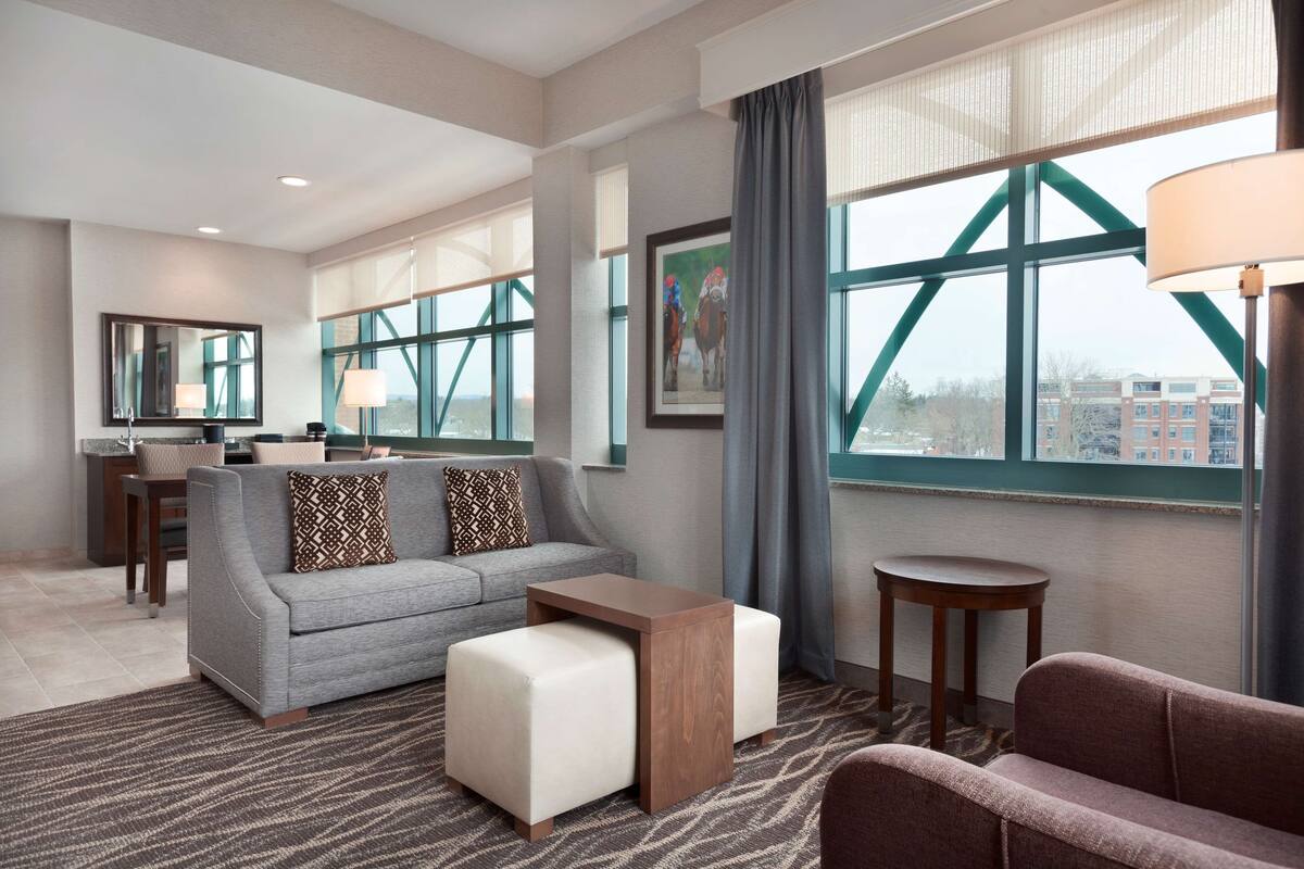 Ein modernes Hotelzimmer mit grauem Sofa, einem Holztisch, einem kleinen runden Tisch und einem großen Fenster mit Blick auf Gebäude.