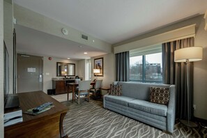 A hotel room with a sofa, a desk, a chair, a lamp, and a window with curtains. - Embassy Suites Saratoga Springs (Saratoga Springs)