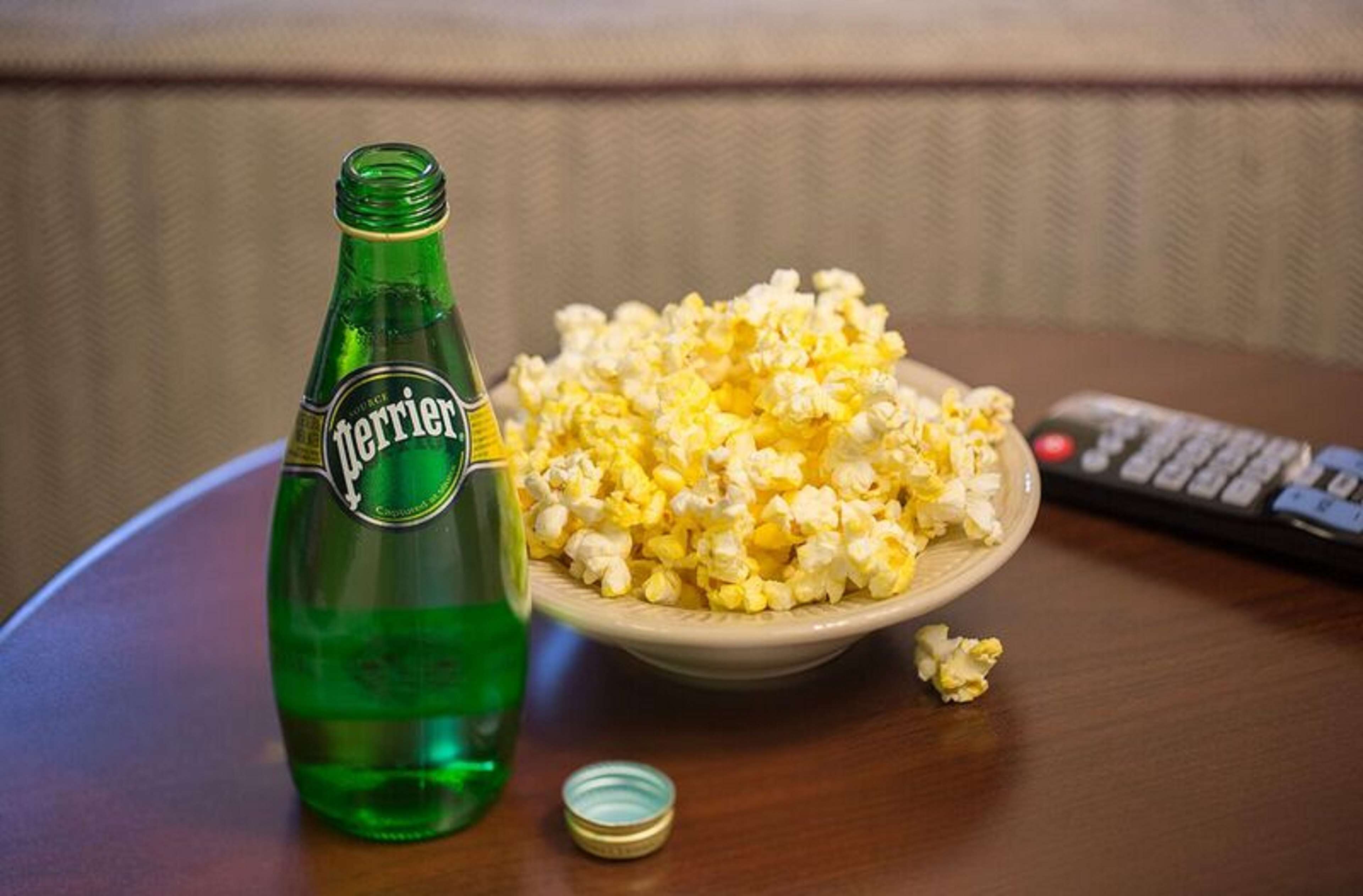 A bottle of Perrier water, a bowl of popcorn, and a remote control on a wooden table.