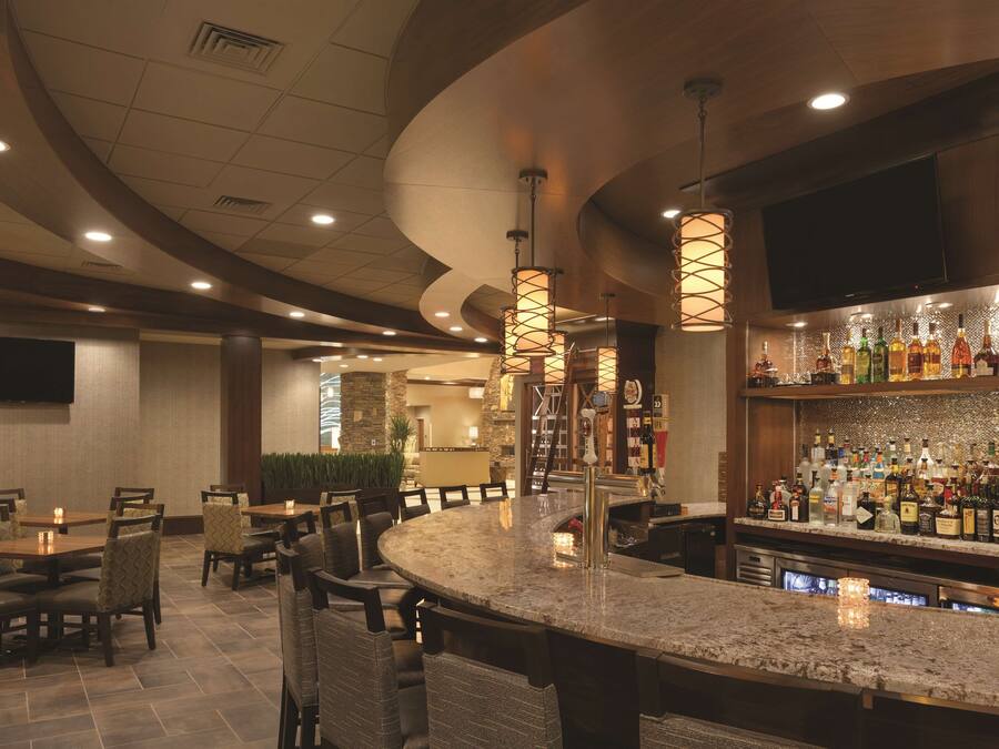 Uma área de bar com bancada de granito curva, diversos arranjos de assentos com cadeiras acolchoadas e uma variedade de garrafas de bebidas alcoólicas nas prateleiras.