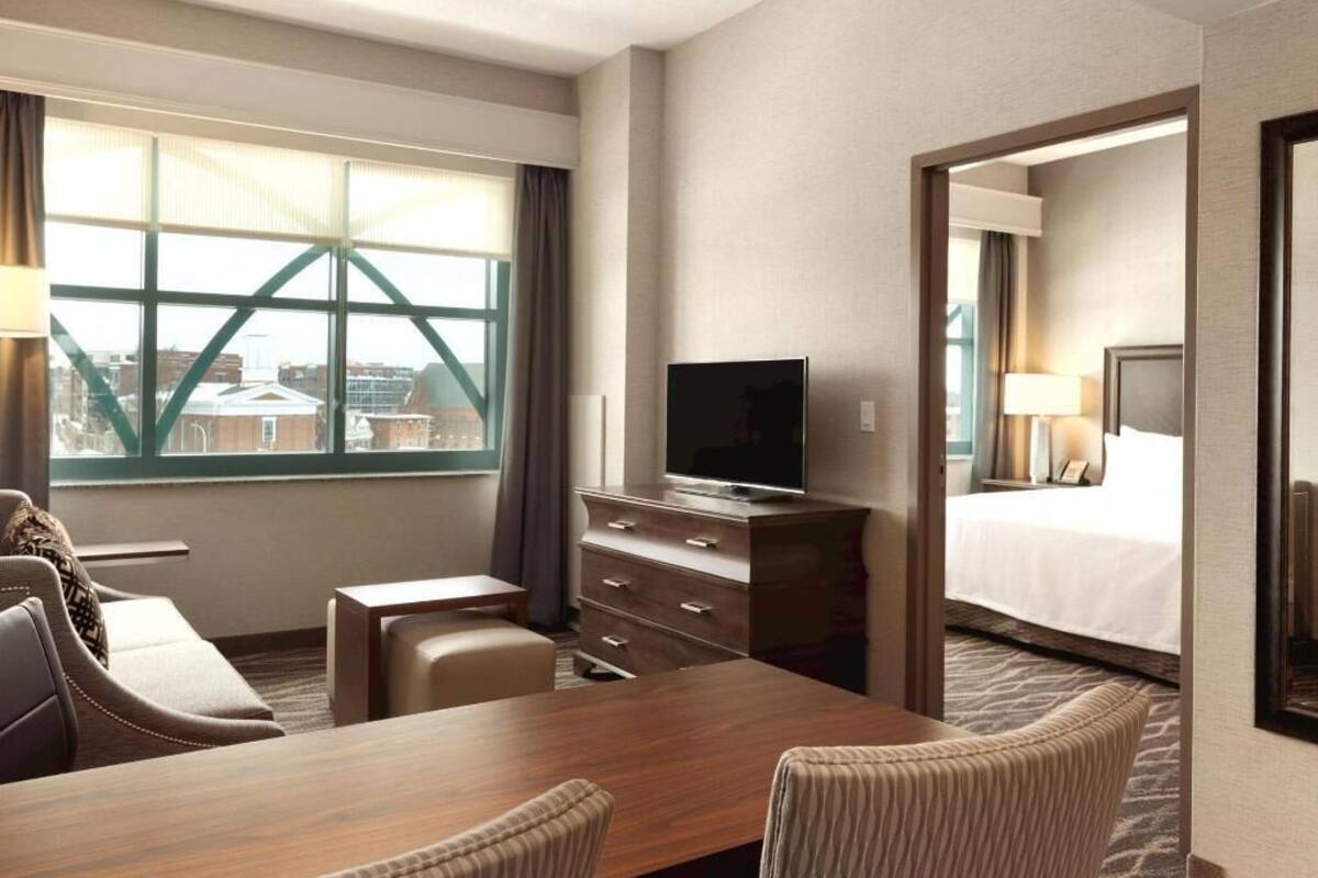 Ein Hotelzimmer mit großem Fenster, Fernseher, Schreibtisch, Stuhl, Kommode und Bett.