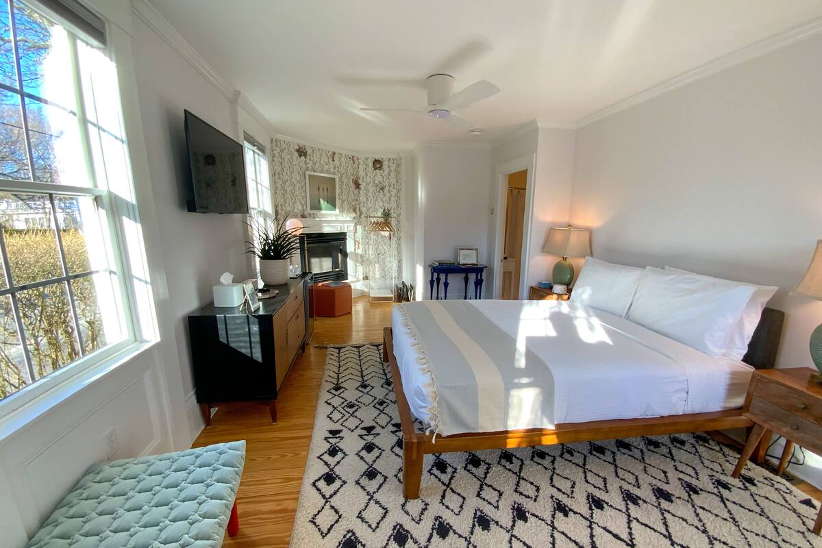 A bedroom with a bed, bedside table, lamp, and a view of the outside.