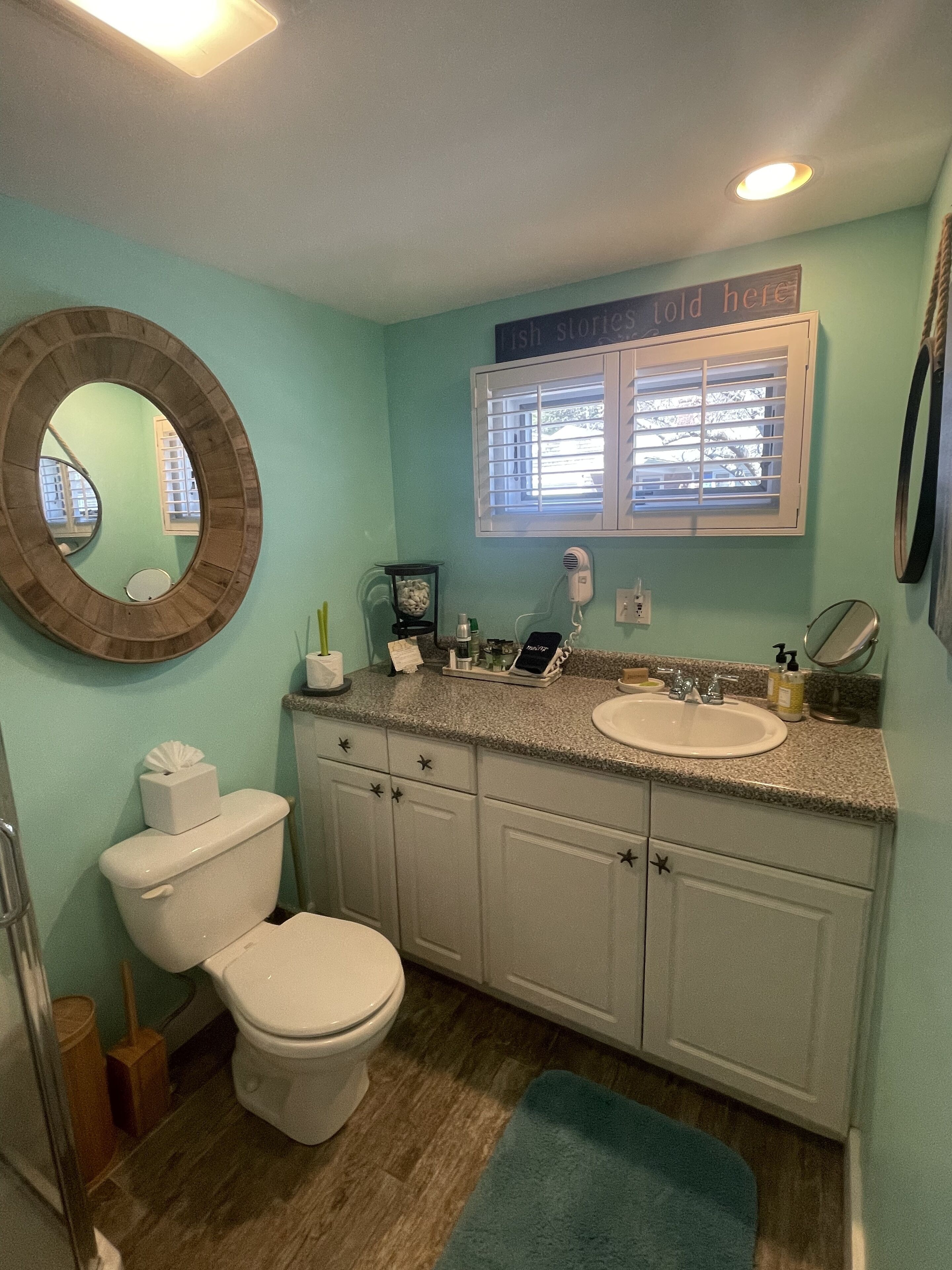 A bathroom with a toilet, sink, and mirror. There is a window with blinds, a wooden-framed mirror, and a sign above the window that says "fish stories here".