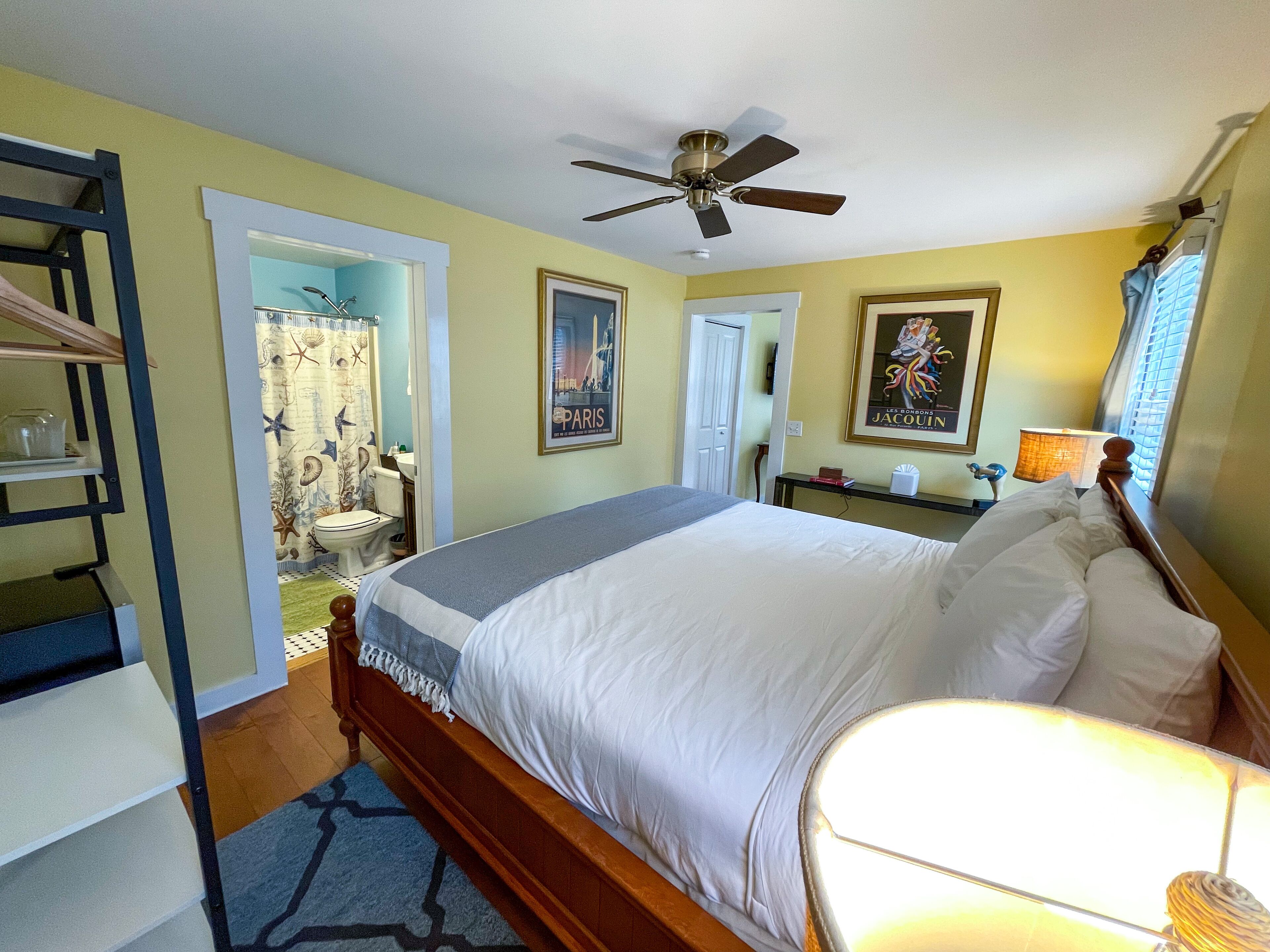 A bedroom with a bed, a ceiling fan, a bathroom with a shower curtain, and a painting on the wall.