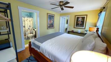 A bedroom with a bed, a ceiling fan, a bathroom with a shower curtain, and a painting on the wall.