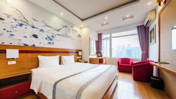 Una habitación de hotel con una cama grande, un cabecero de madera, un sillón rojo, un escritorio y un ventanal con cortinas.