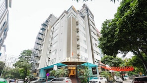 Un hotel de varios pisos con una entrada destacada, una escalera que conduce al vestíbulo y una calle urbana circundante con coches aparcados y árboles.