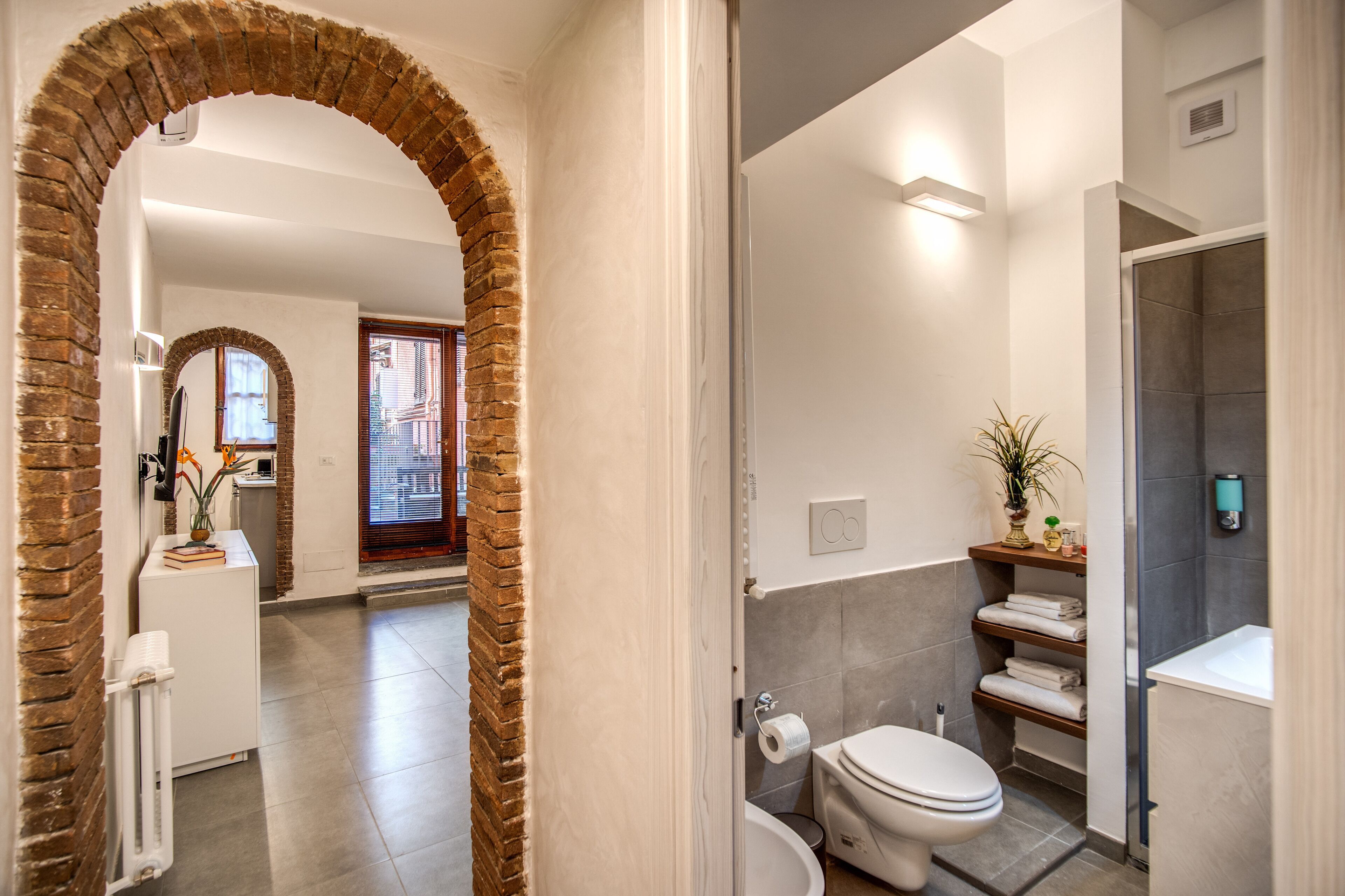 Een moderne badkamer met toilet, wastafel en douche. Een bakstenen boog leidt naar een andere ruimte.