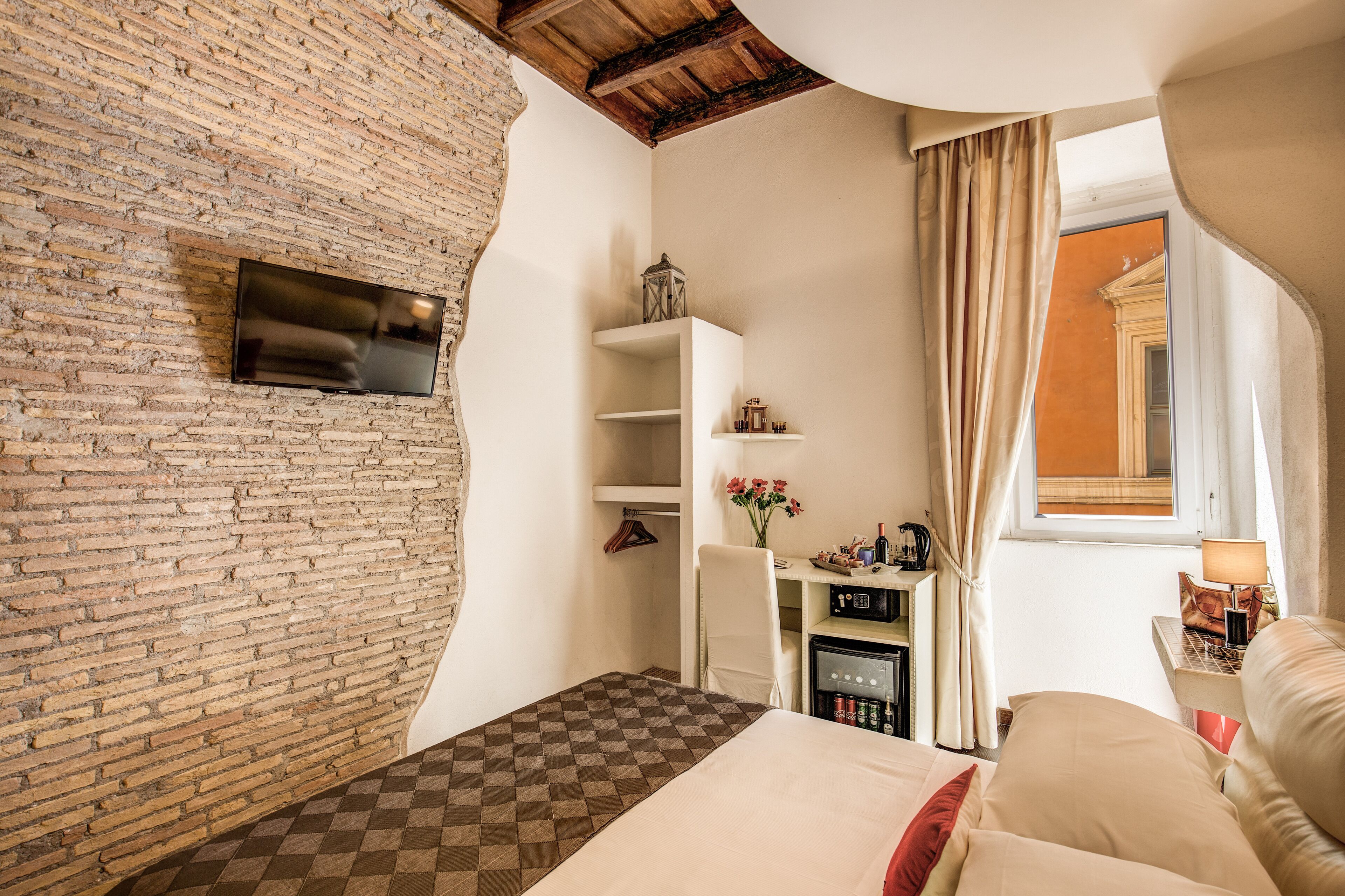A bedroom with a brick wall, a bed with a checkered bedspread, a white wardrobe, a small table with a lamp, and a window with curtains.