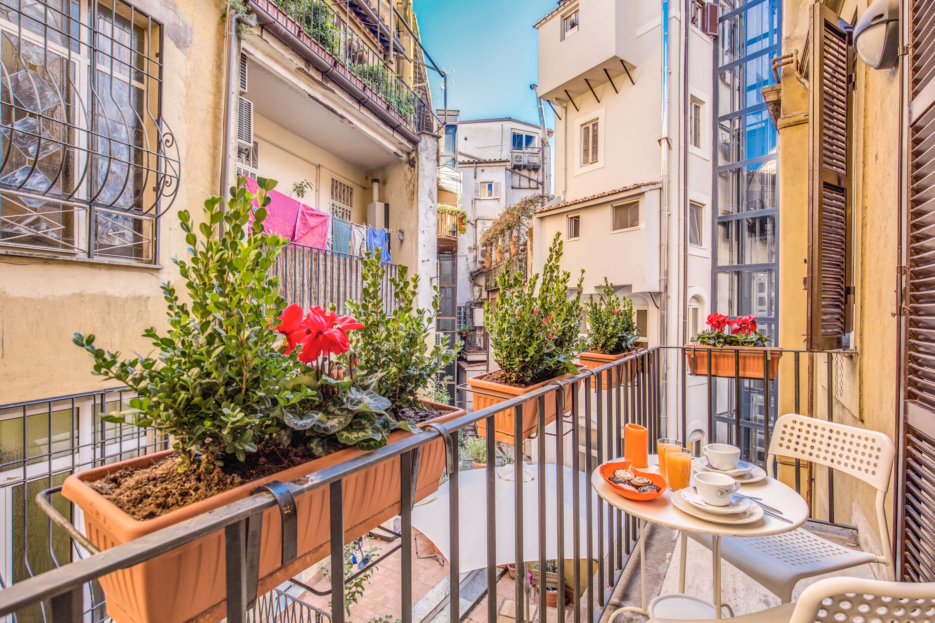 A narrow urban street with balconies, potted plants, and outdoor seating.