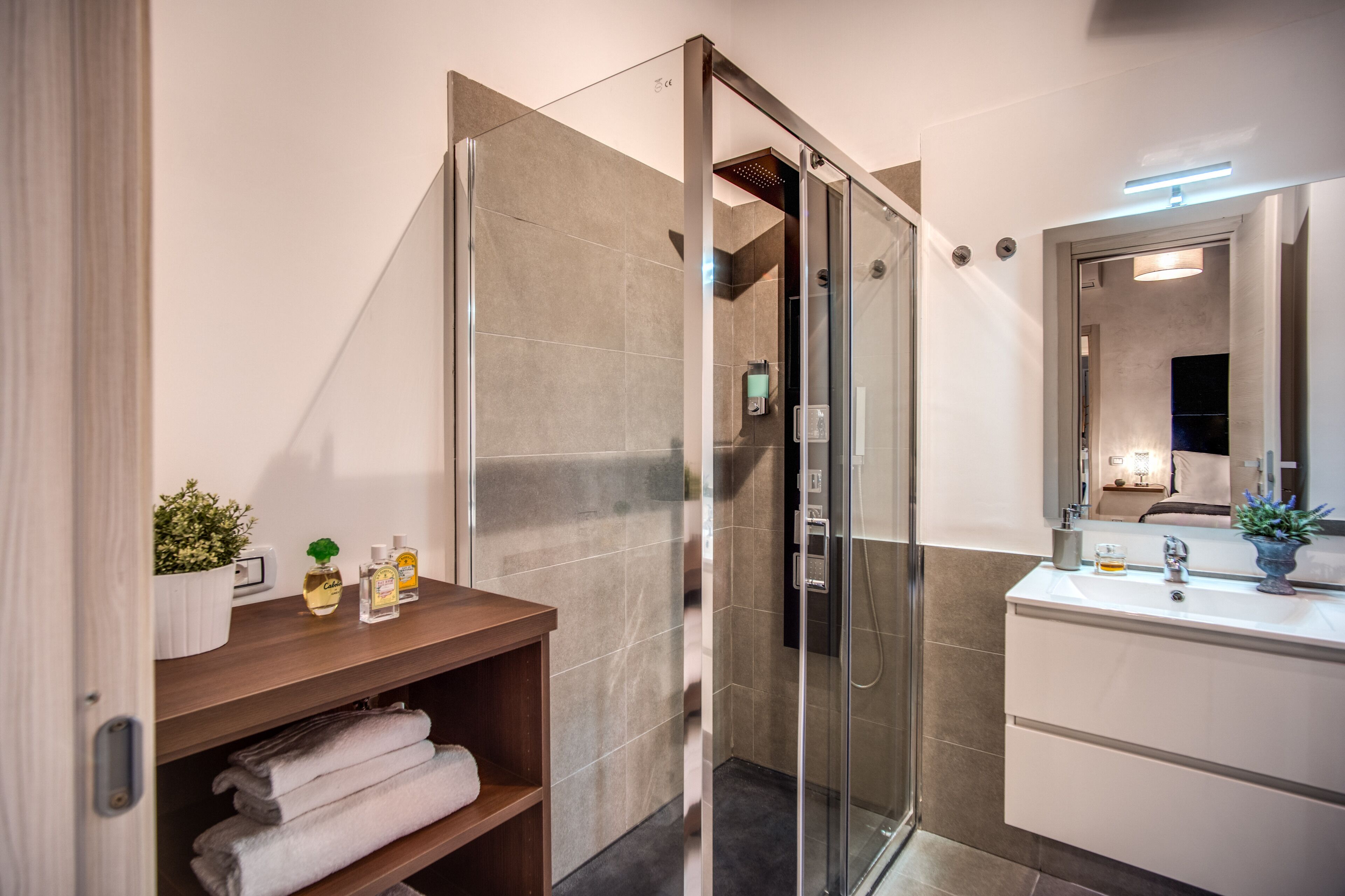 Une salle de bain moderne avec une douche à parois de verre, une étagère en bois avec des serviettes, un lavabo blanc et un miroir avec un éclairage au-dessus.