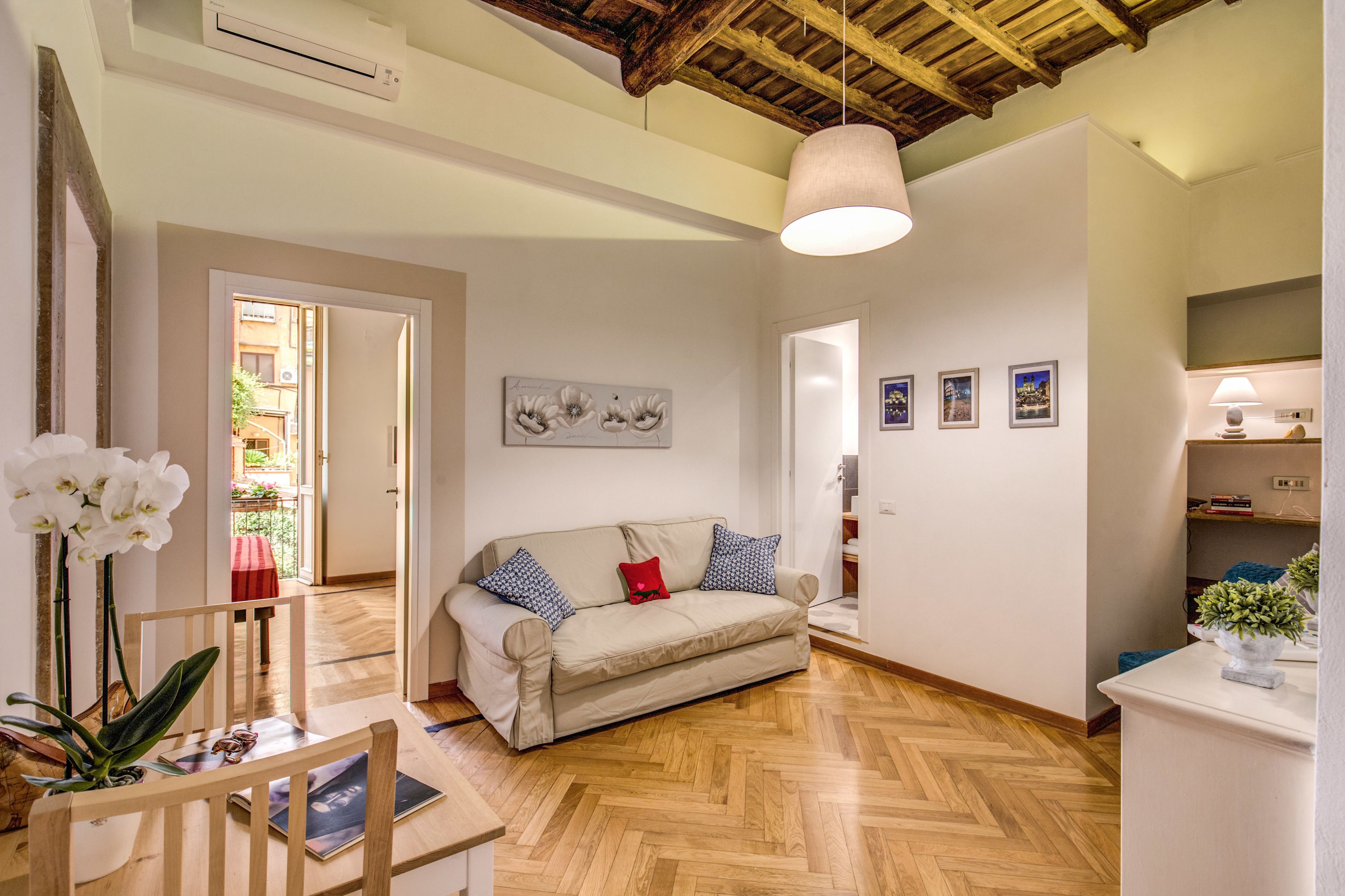 A spacious living room with a wooden herringbone floor, a beige sofa with patterned cushions, a wooden coffee table, and a hanging light fixture.