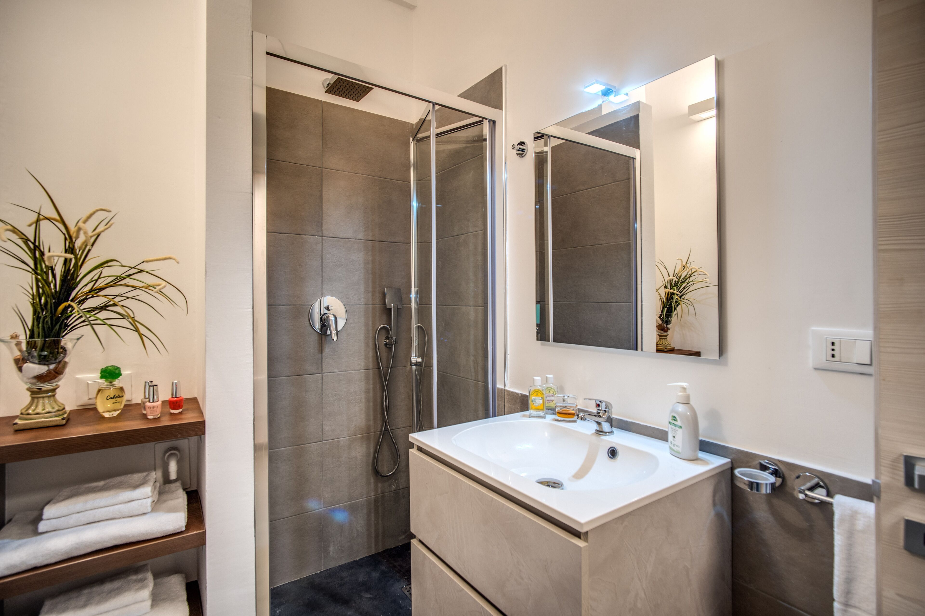 A modern bathroom with a glass-enclosed shower, a white sink, and a mirror above it. There are towels on a shelf and a potted plant on the left.