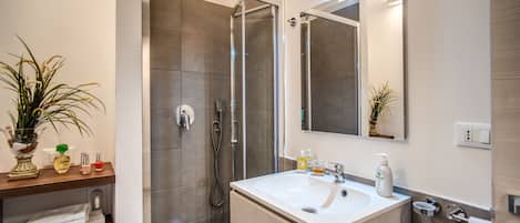 A modern bathroom with a glass-enclosed shower, a white sink, and a mirror above it. There are towels on a shelf and a potted plant on the left.