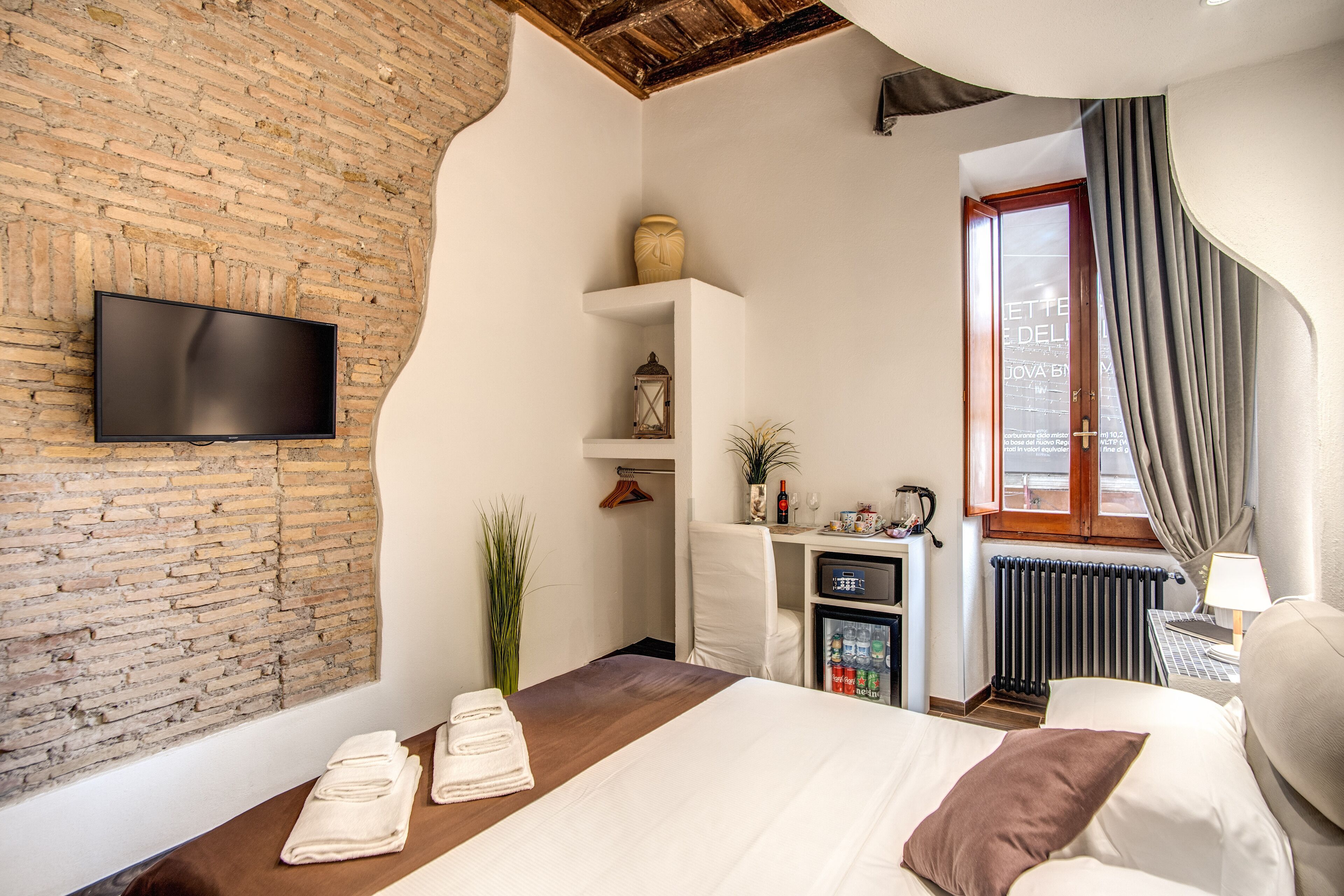 Quarto de hotel com parede de tijolos, televisão de tela plana fixada nela, cama com roupa de cama branca e travesseiro marrom, mesa pequena com toalhas, radiador, escrivaninha com computador, planta e janela com cortinas.