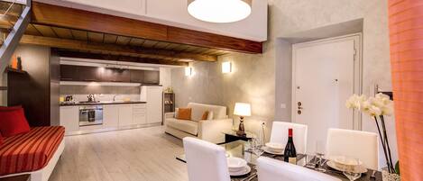 A modern living room with a sofa, dining table, and chairs, connected to a kitchen area with a built-in oven and sink.