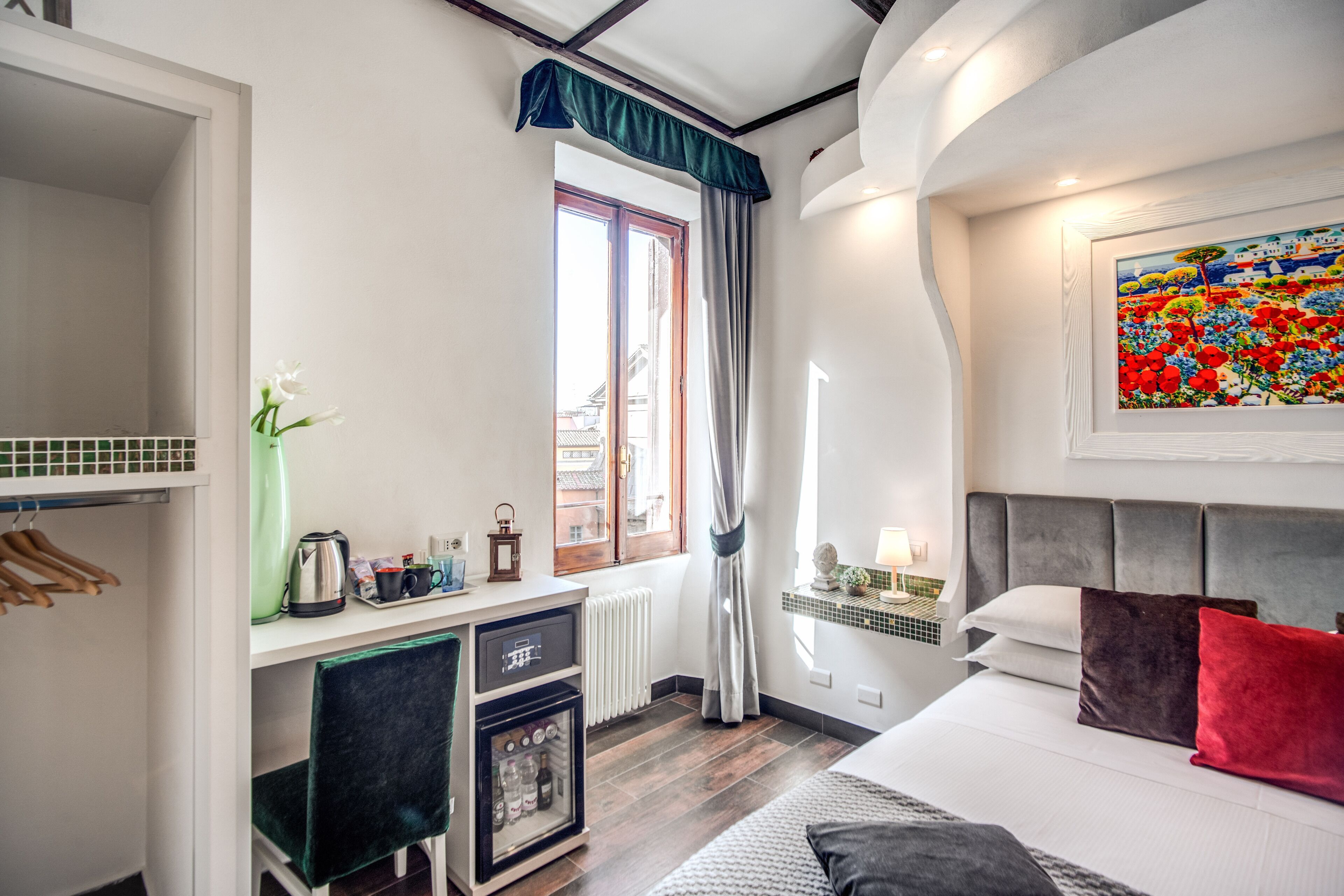 A hotel room with a bed, a desk with a kettle, a radiator, and a window with curtains.