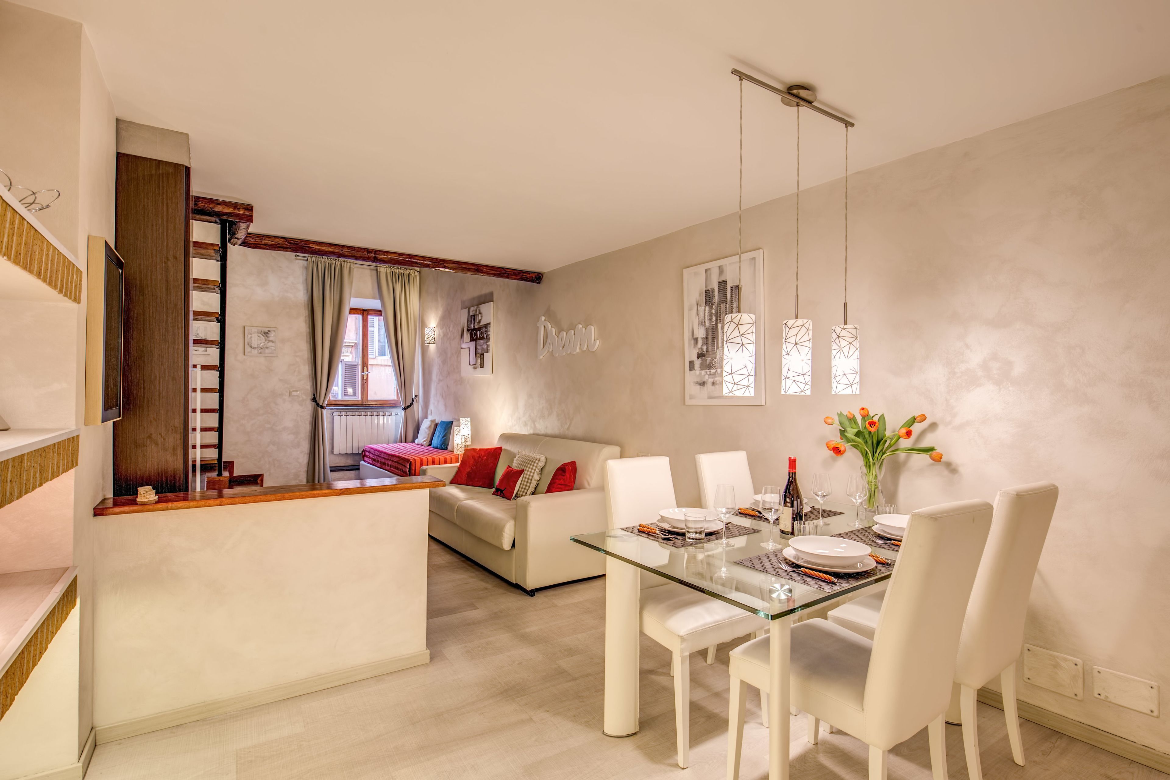 A modern living room with a glass dining table, white chairs, a sofa with red cushions, a staircase, and a cityscape artwork on the wall.