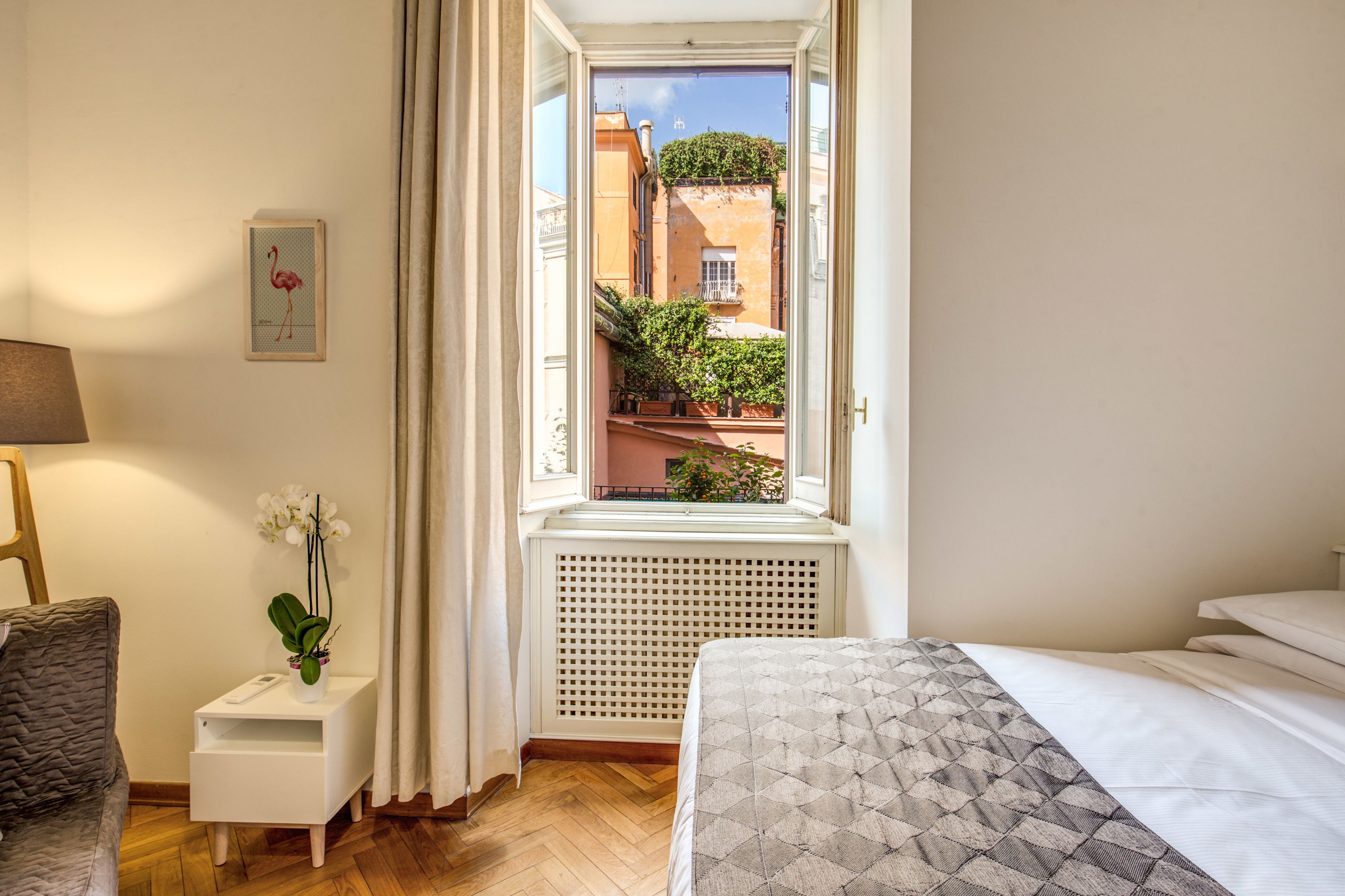 A hotel room with a bed, a small bedside table with a plant, a window with a view of the city, and a framed picture on the wall.