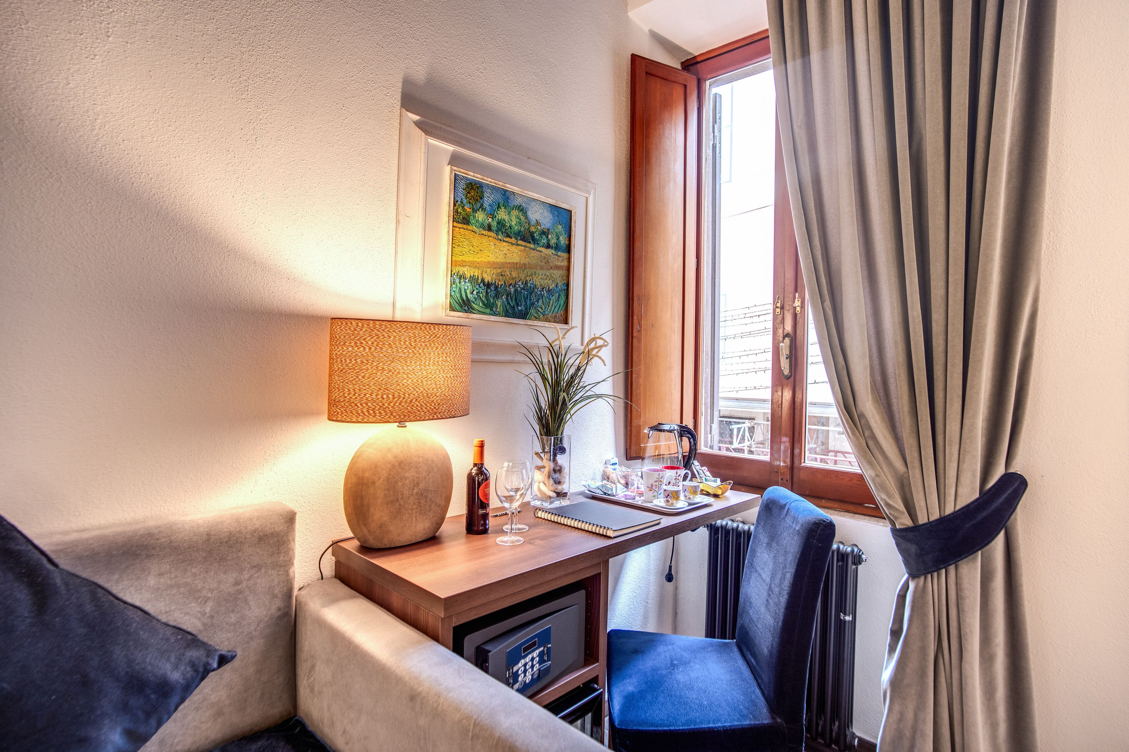 Una habitación de hotel acogedora con un escritorio de madera, una silla cómoda, una lámpara, un cuadro y vistas de la ciudad a través de una ventana con cortinas.