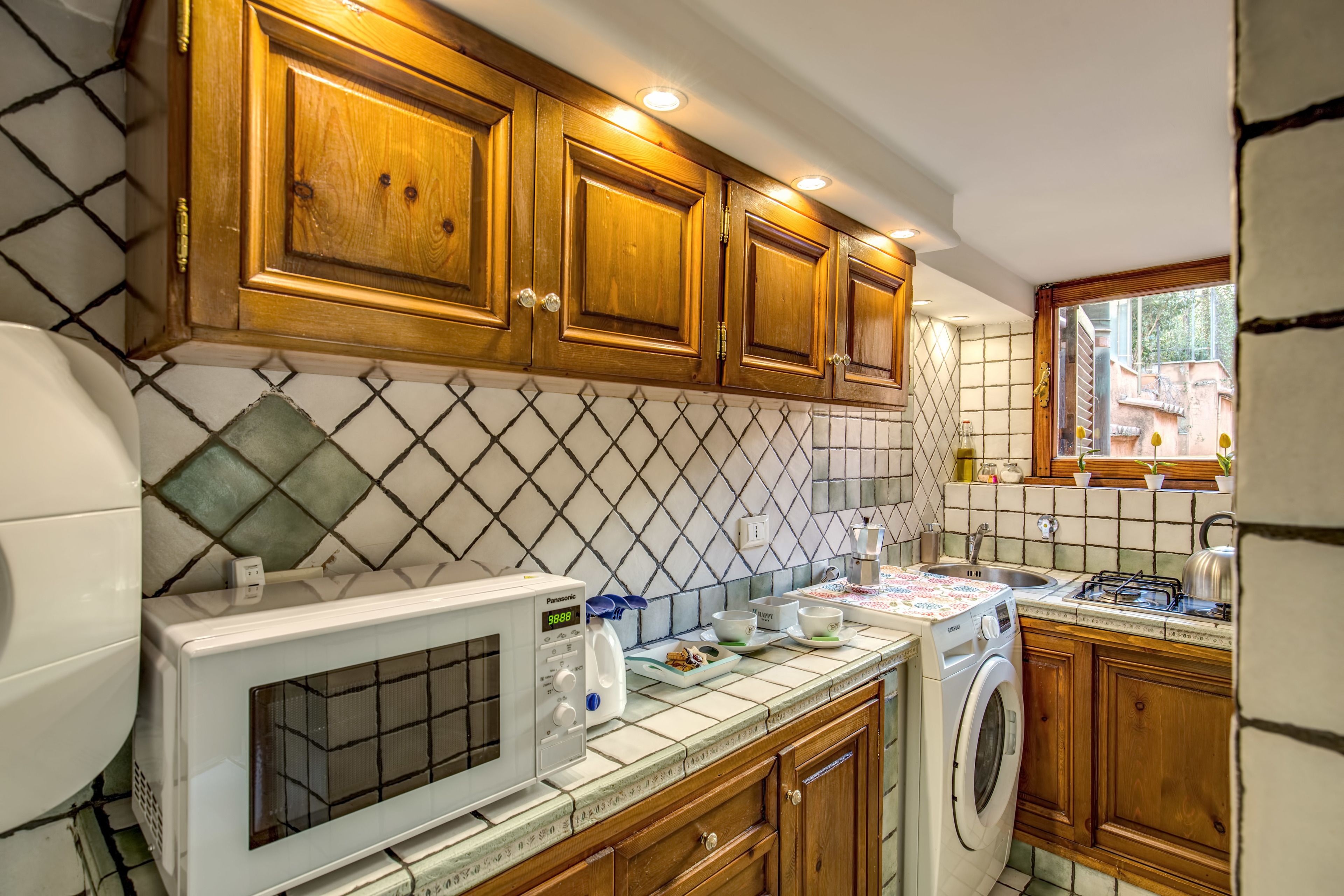 Cucina con mobili in legno, forno a microonde, lavatrice e parete di fondo rivestita di piastrelle.