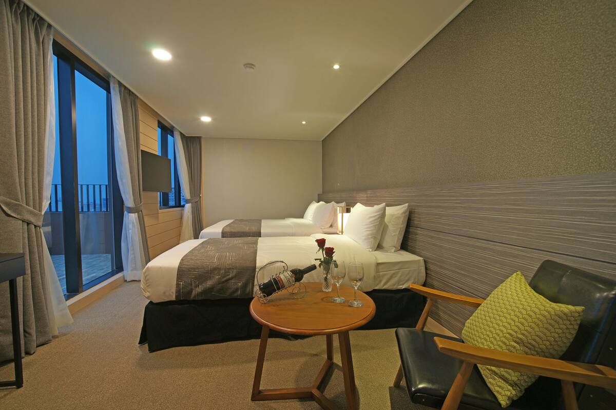 Habitación de hotel con cama, una pequeña mesa con una botella y vasos, una silla y vistas a la ciudad a través de amplias ventanas.