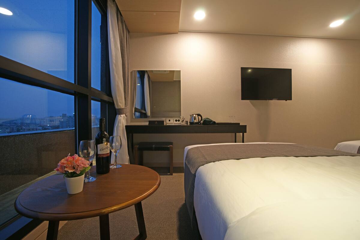 Habitación de hotel con una cama, una mesita con una botella y vasos, un televisor montado en la pared y vistas a la ciudad a través de la ventana.