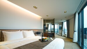 Habitación de hotel con una cama grande, una mesita de noche con una botella y flores, un escritorio con una computadora y ventanas amplias que ofrecen vistas.