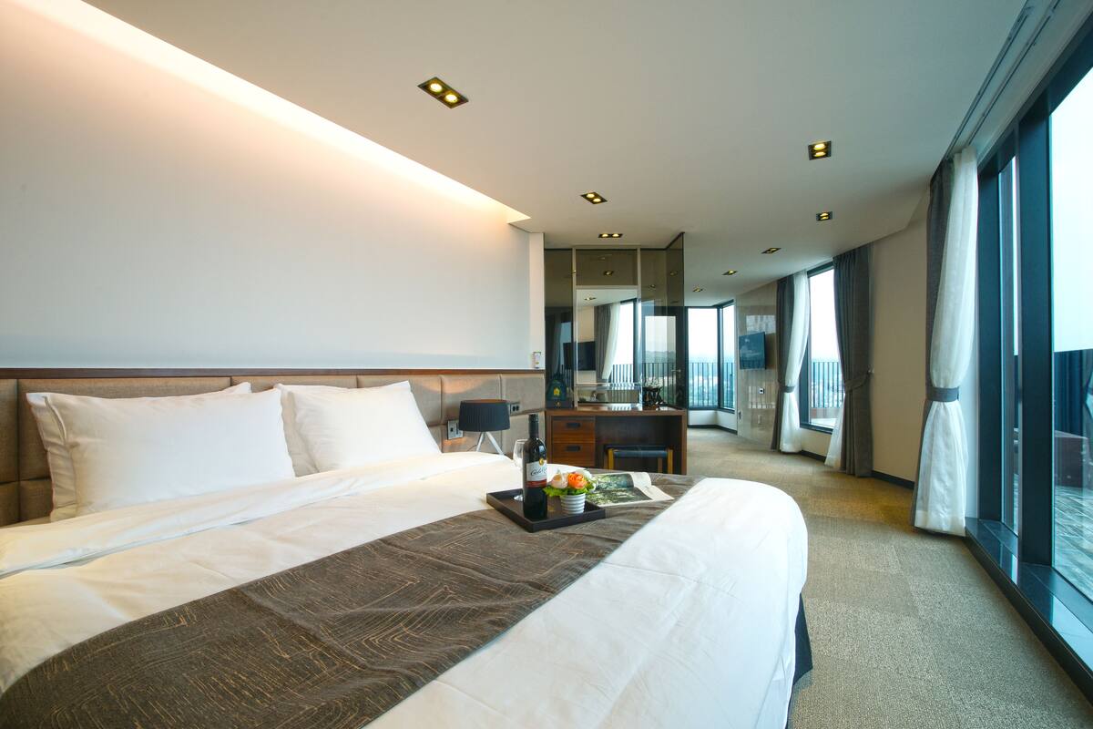 Habitación de hotel con una cama grande, una mesita de noche con una botella y flores, un escritorio con una computadora y ventanas amplias que ofrecen vistas.